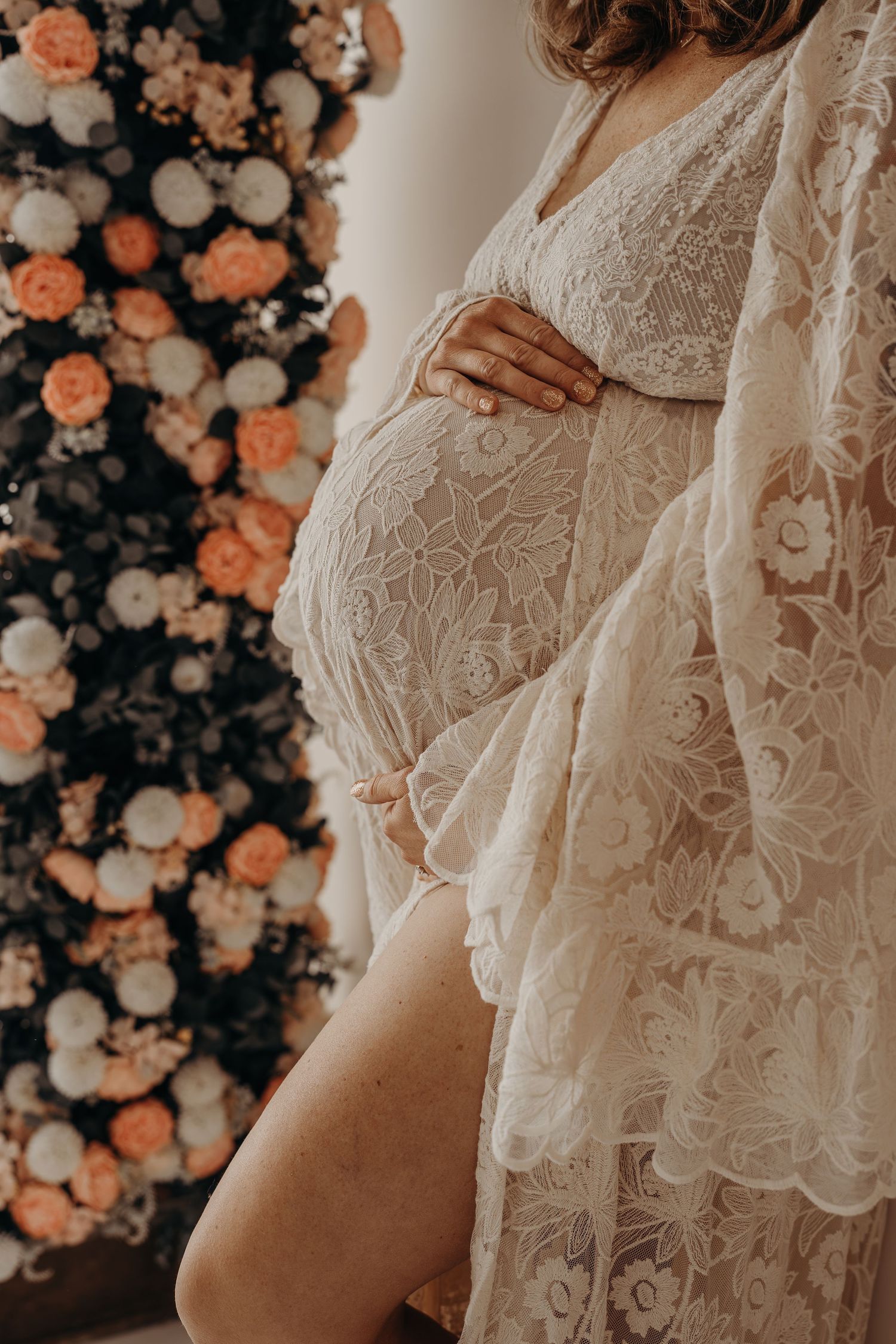 Pregnant woman in white lace maternity gown cradling her baby bump, standing before a floral backdrop during a motherhood mini session.