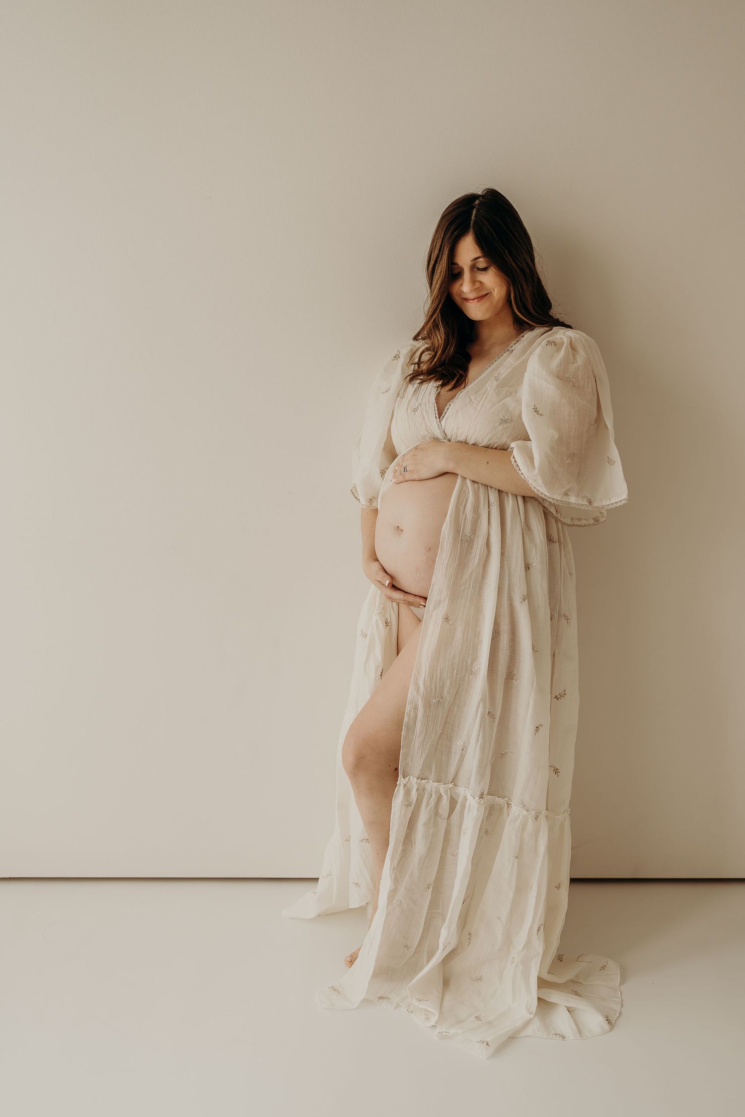 Romantic maternity portraits in cream-colored flowing dress against neutral studio background.
