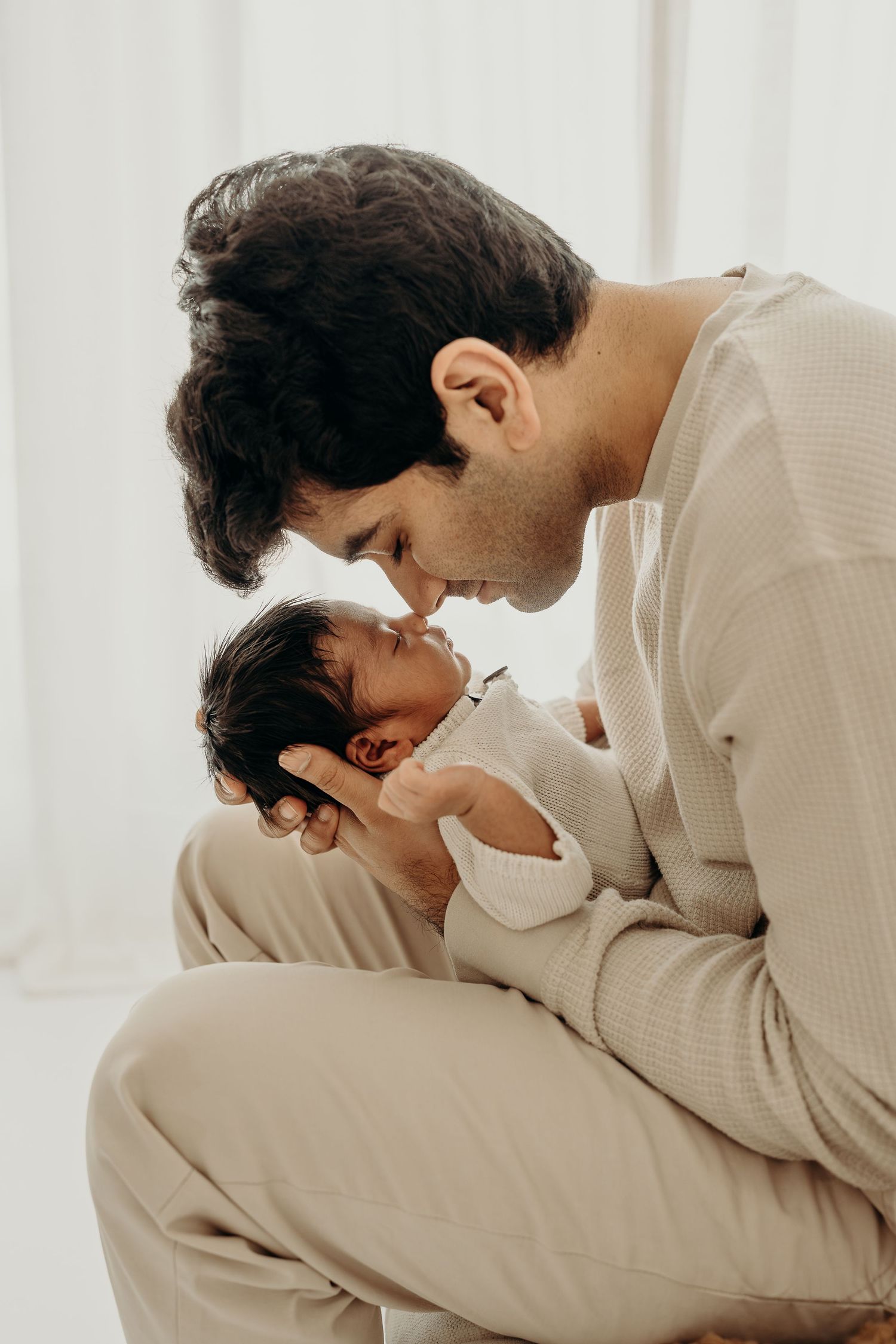 A father tenderly kisses his newborn baby while cradling them gently in a soft, neutral-toned setting.