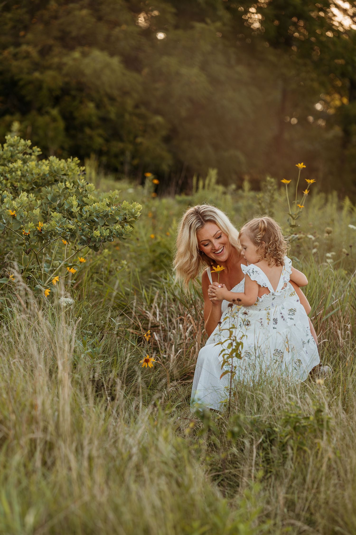 A tender moment shared between two people in white dresses sitting among tall wildflowers at sunset.