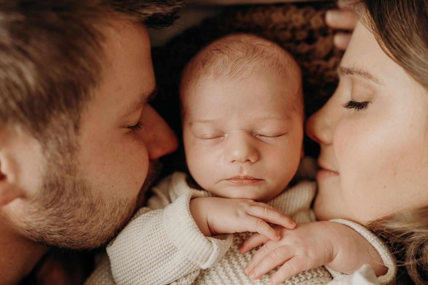 Parents tenderly kissing their sleeping newborn baby, captured in a warm and intimate family portrait.