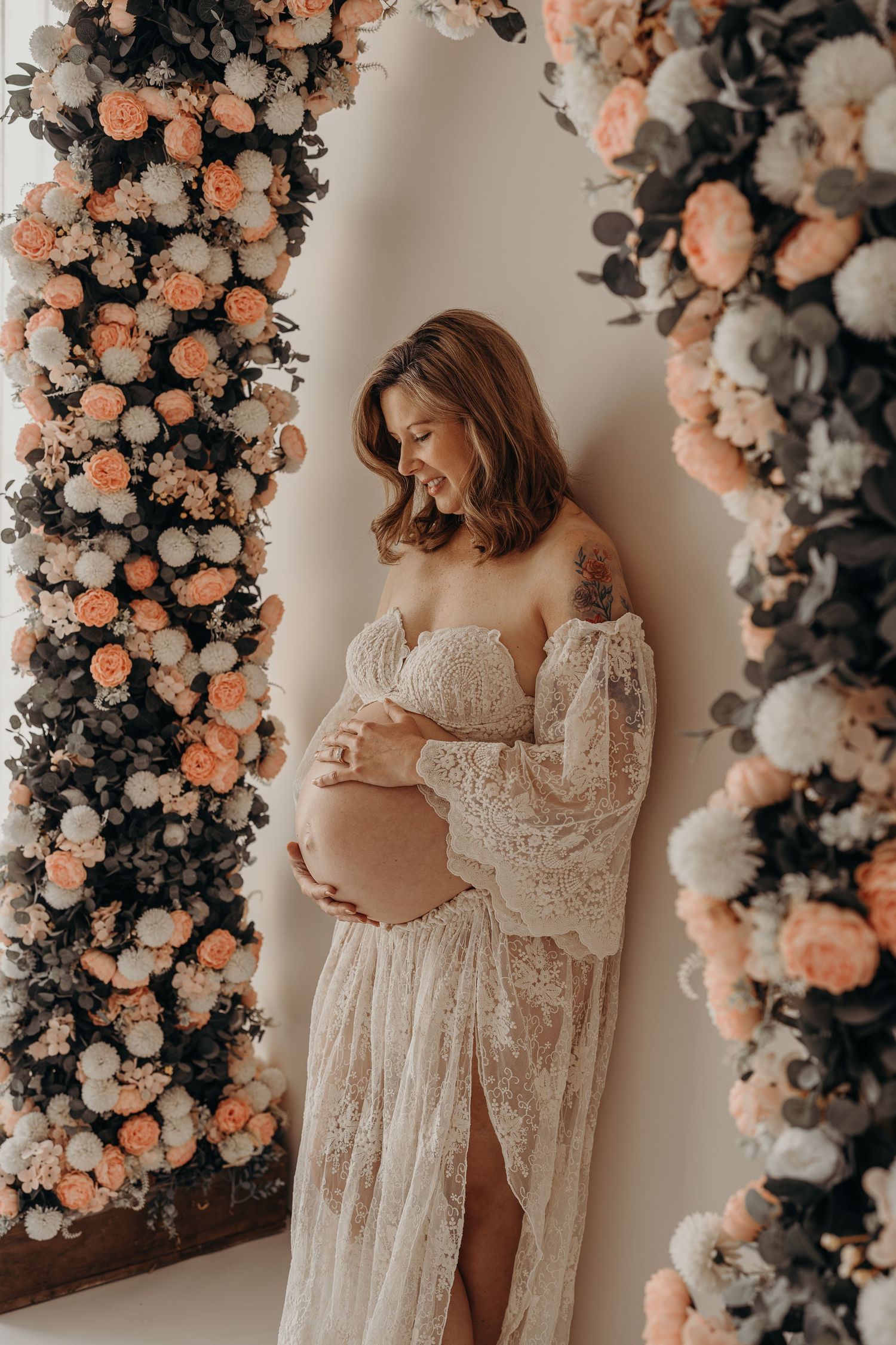 Pregnant woman in white lace gown cradling baby bump under floral arch with peach and white roses during a motherhood mini photo session.