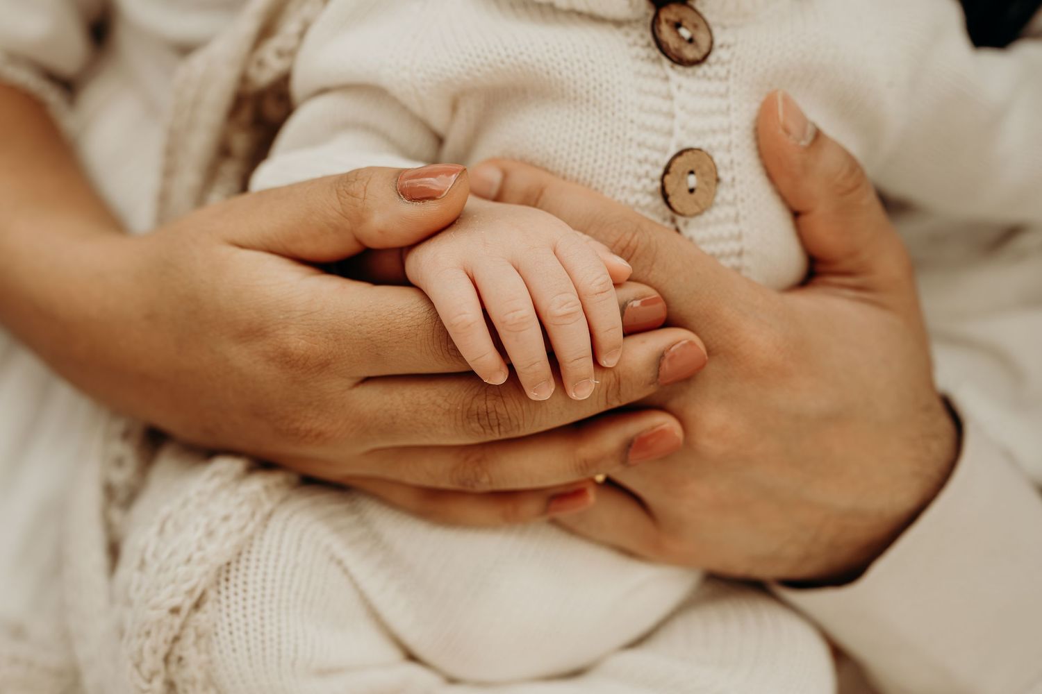 Adult hands tenderly cradling a newborn baby's tiny hand, dressed in cream knit clothing.