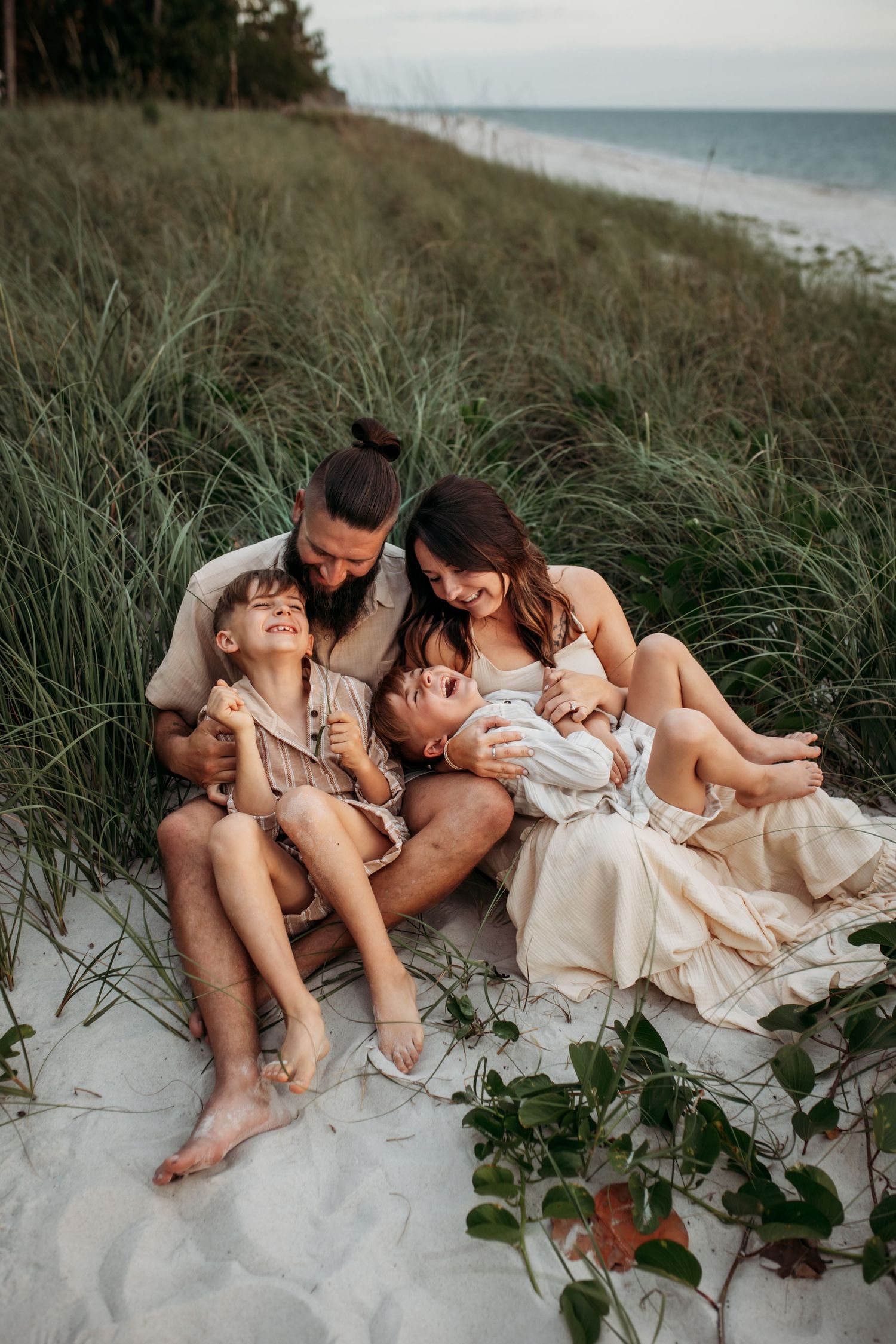 A joyful family of four sits together on white sand dunes surrounded by tall beach grass, sharing laughter and cuddles.