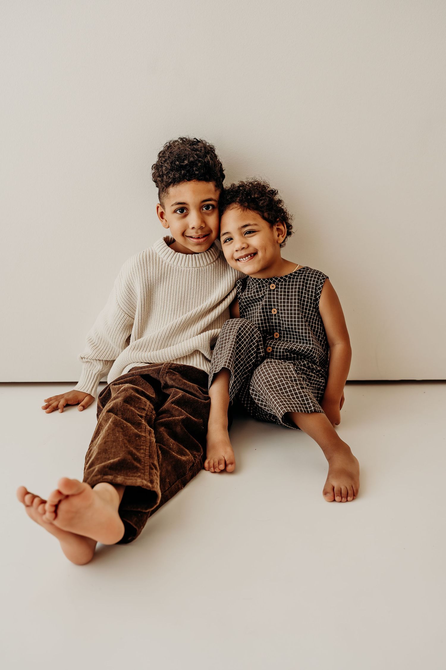 Two smiling children sitting together on floor, boy in cream sweater and brown pants, girl in checkered jumper.