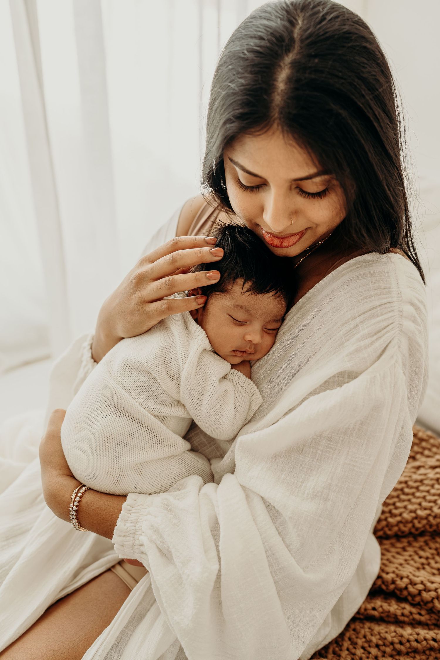 A mother tenderly cradles her sleeping newborn baby, both dressed in soft white, in a warm, serene setting.