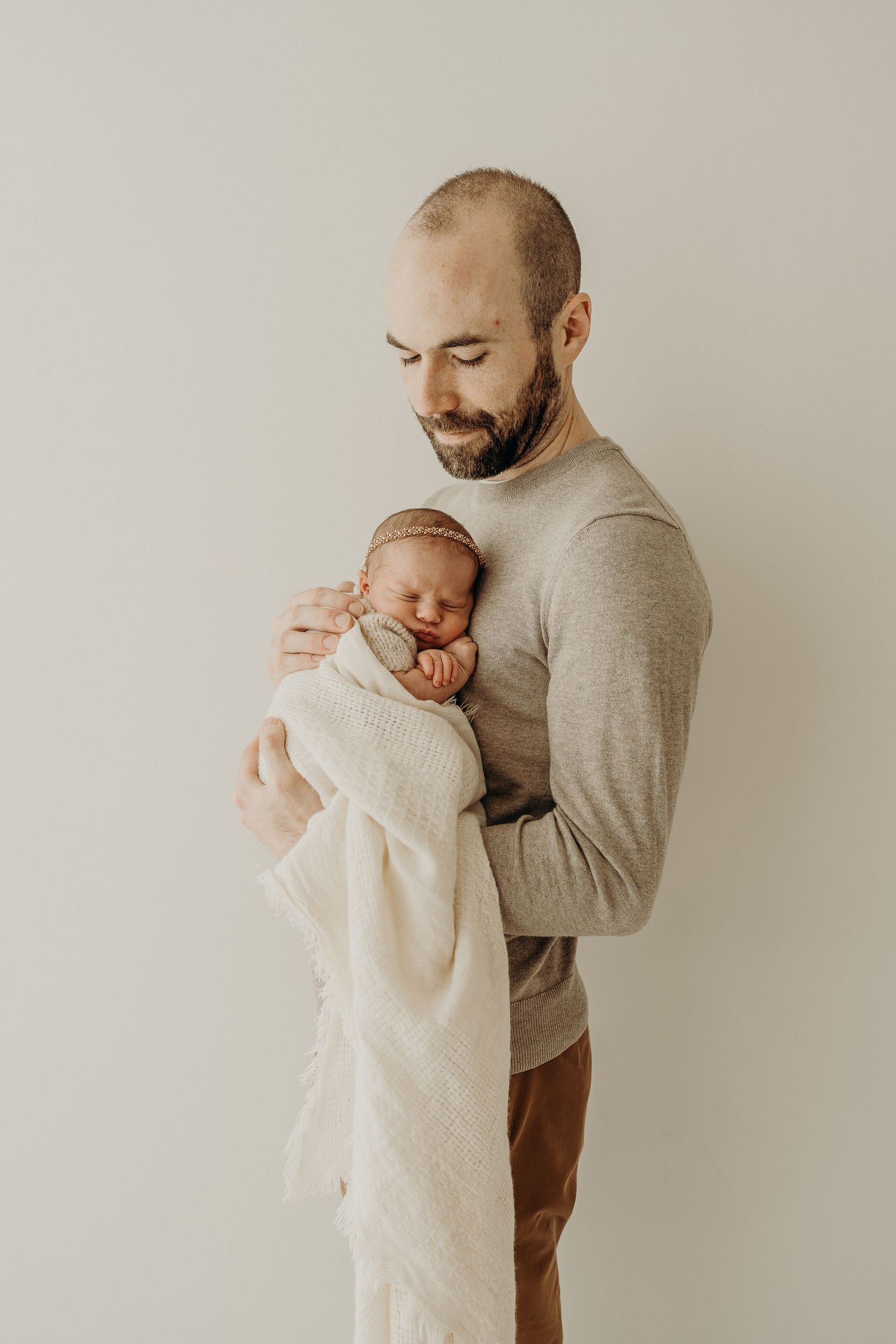 A new father in a beige sweater tenderly holds a sleeping newborn wrapped in a cream blanket, during a newborn photography session