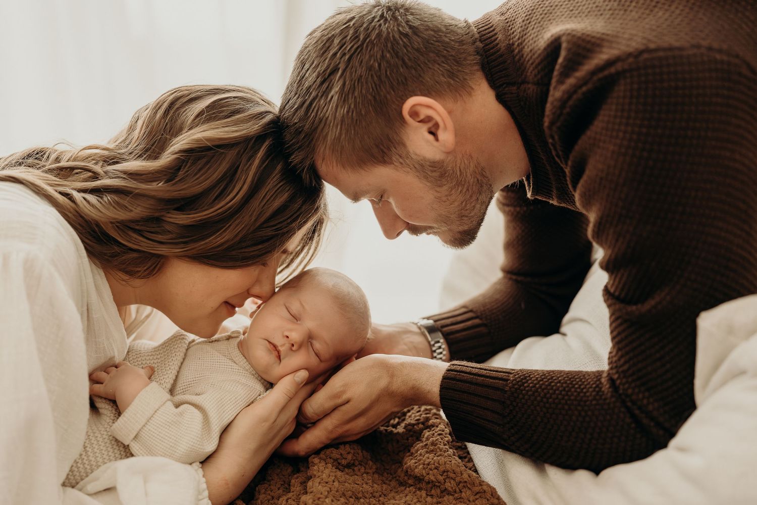 New parents lovingly gaze at their sleeping newborn baby wrapped in a cozy brown blanket.