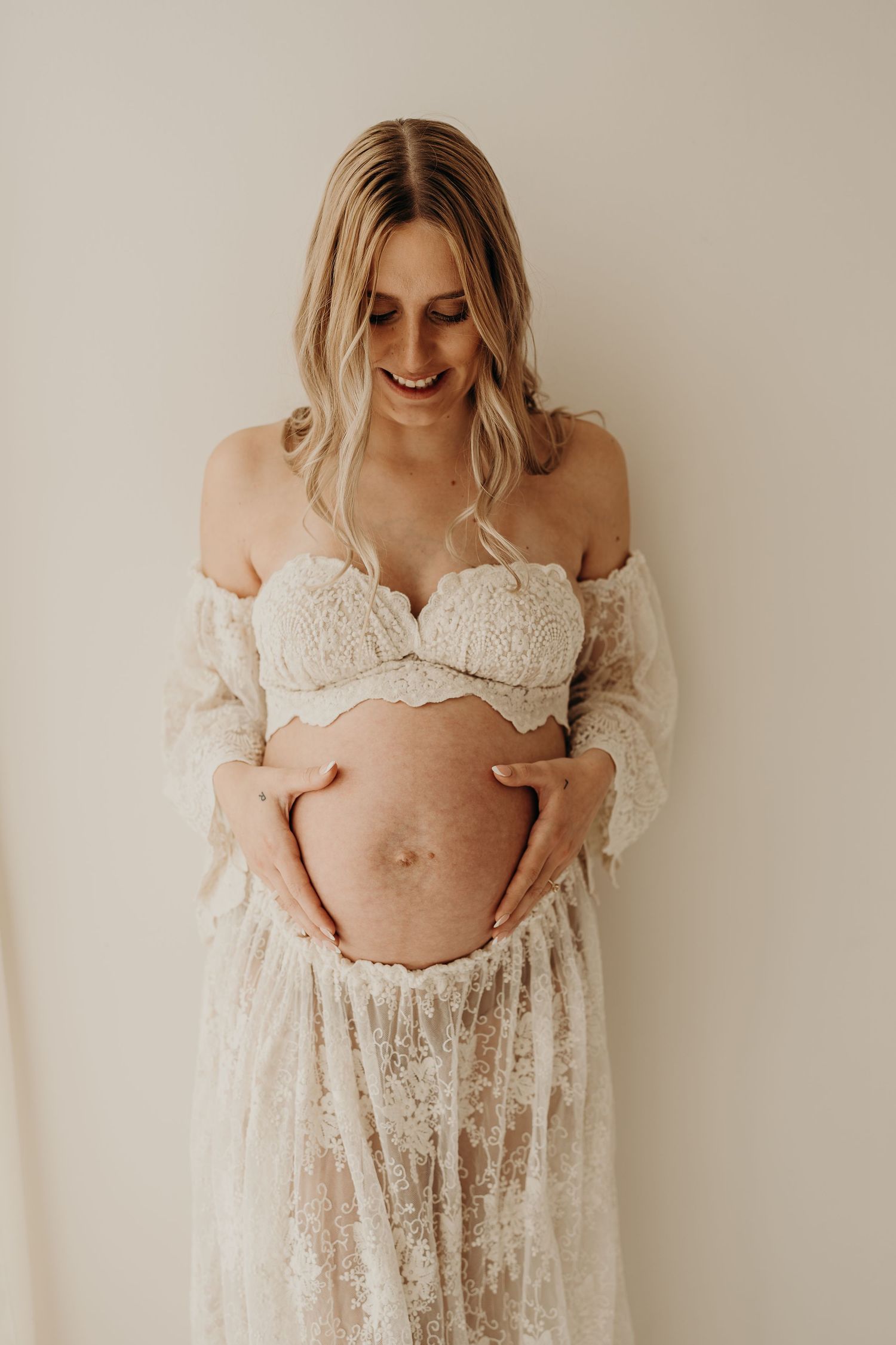 A pregnant woman in a delicate white lace maternity gown poses against a neutral background in a series of photos.