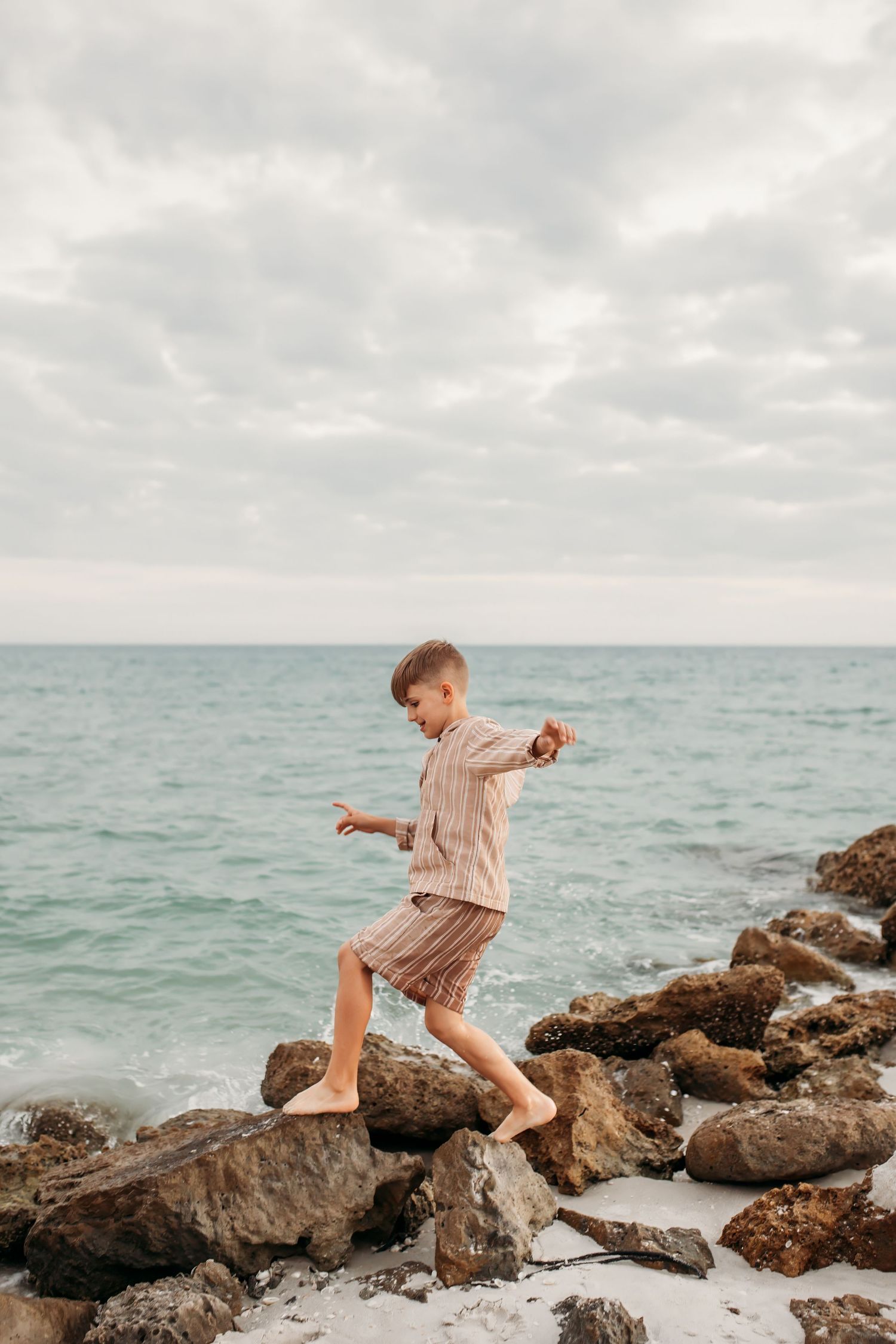 A barefoot boy carefully balances on rocky shoreline with turquoise ocean waves and cloudy sky in background.