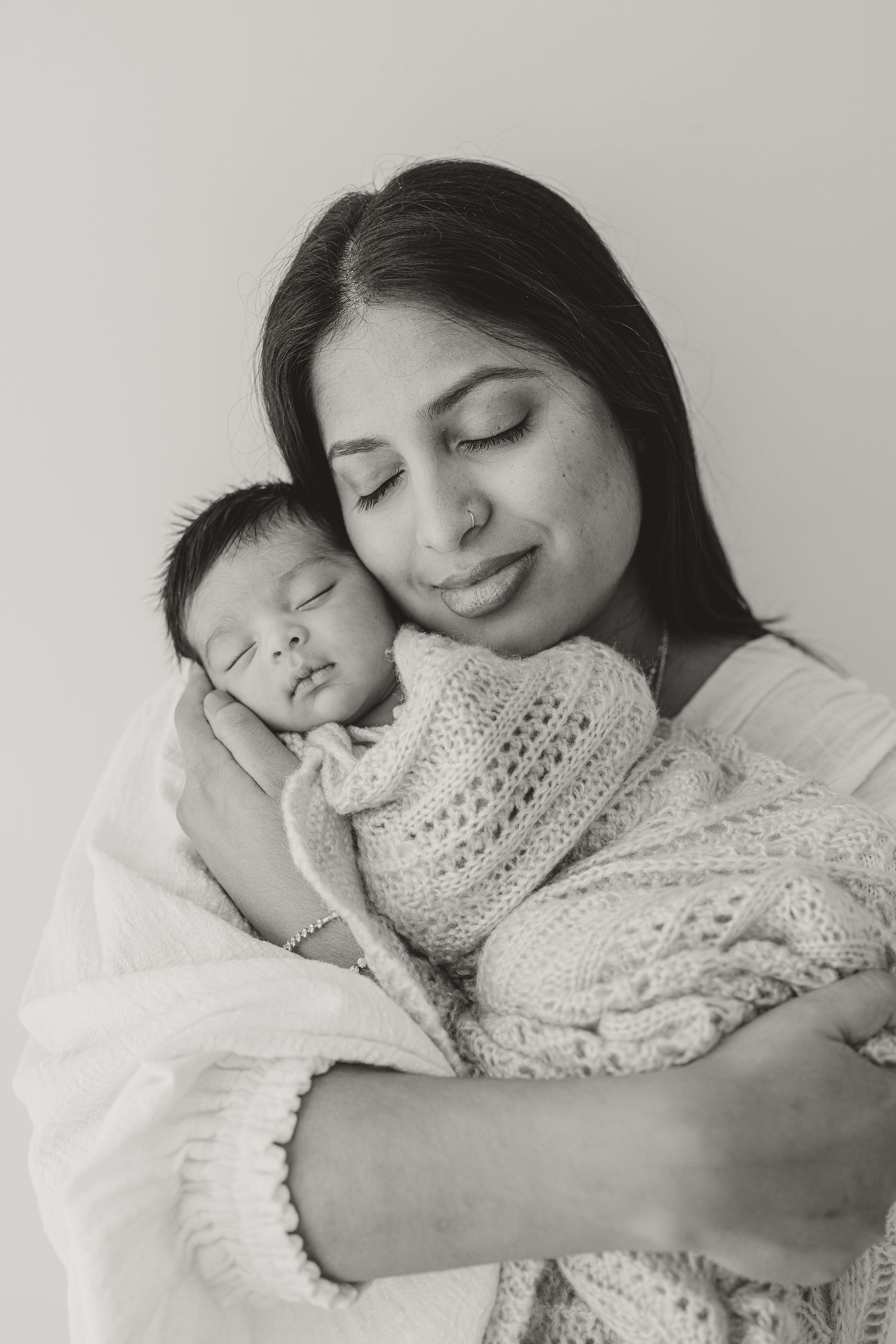 Black and white photo of a mother tenderly holding her sleeping newborn baby wrapped in a knit blanket.