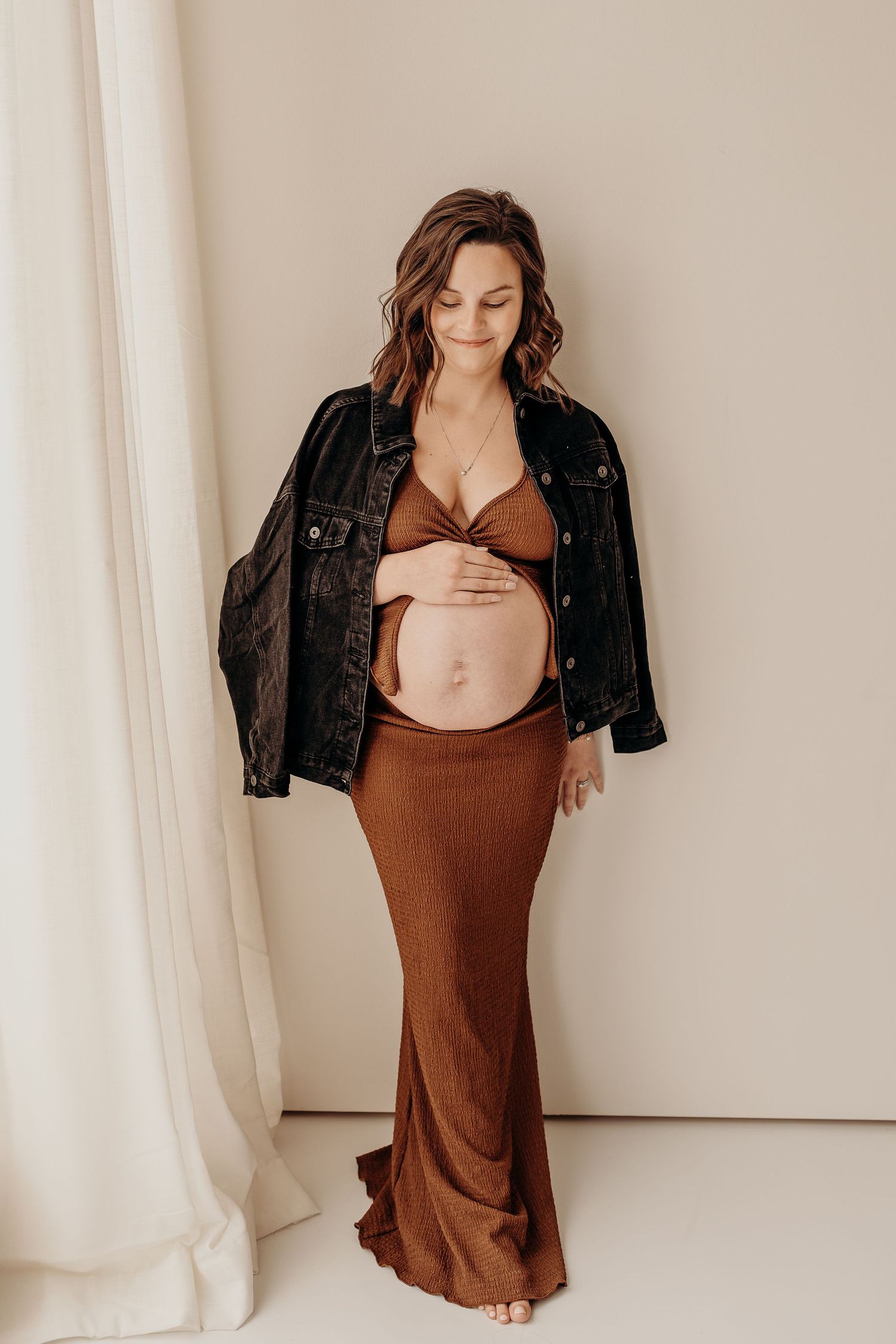 Model in brown slip dress and black leather jacket poses against white backdrop in artistic fashion series.