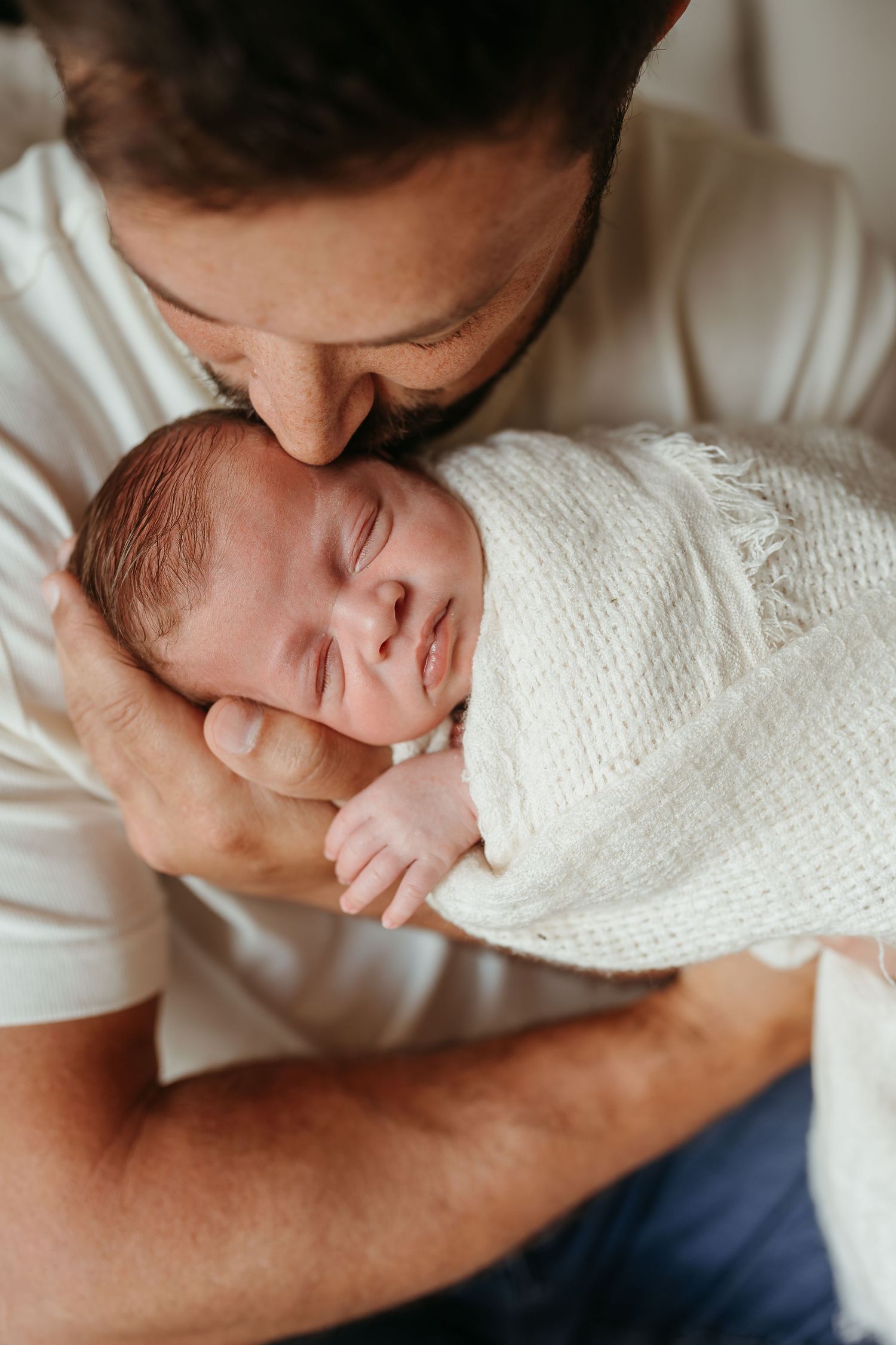 Tender moments of a newborn baby being held and kissed while wrapped in a white knit blanket.