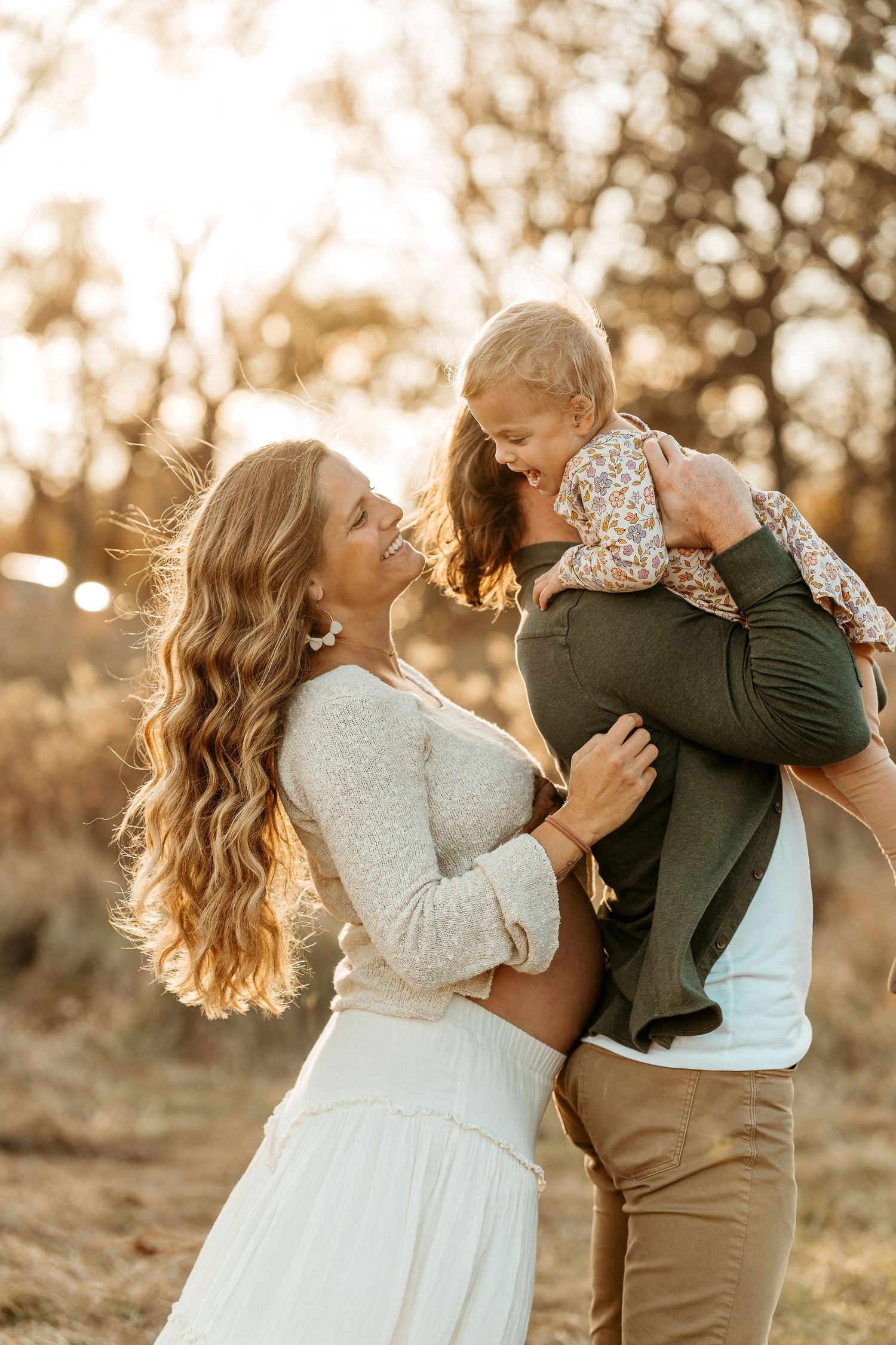 Joyful family moment at sunset with parents lifting child in golden outdoor lighting showcasing maternity and motherhood journeys.