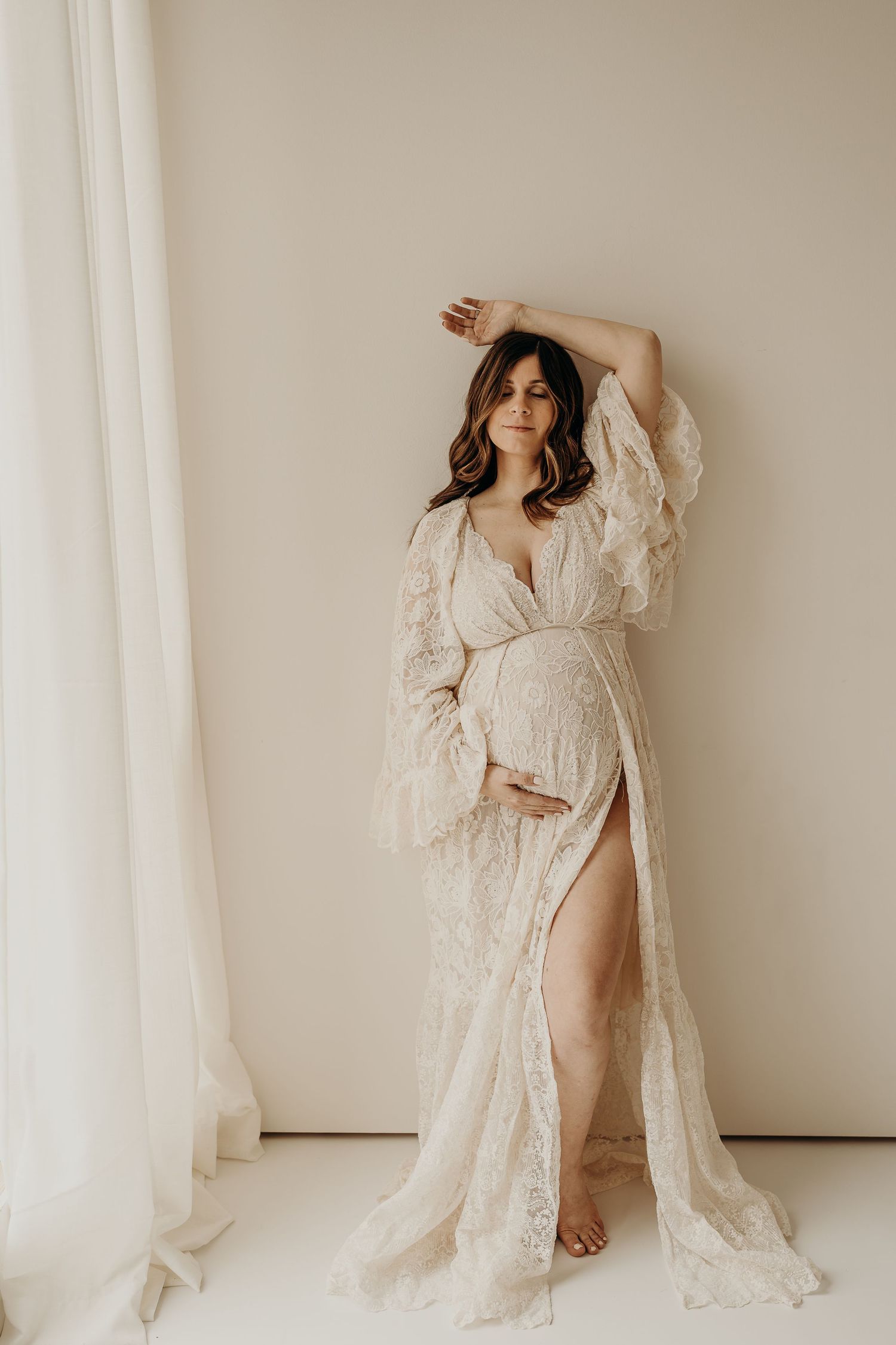 Ethereal maternity photoshoot featuring a woman in flowing white lace robe against neutral backdrop.