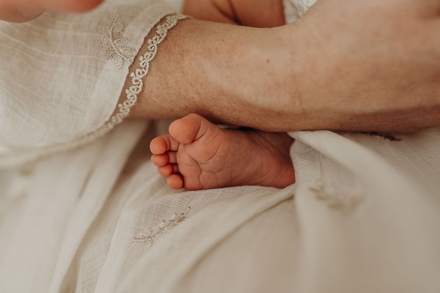 Close-up of a newborn baby's tiny feet resting on a soft white blanket, gently held by an adult's hand.