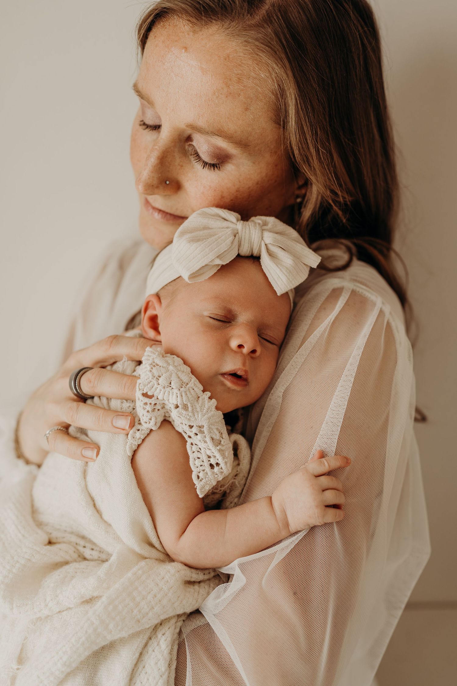 A freckled mother tenderly holds her sleeping newborn baby dressed in a cream crochet outfit with a bow headband.