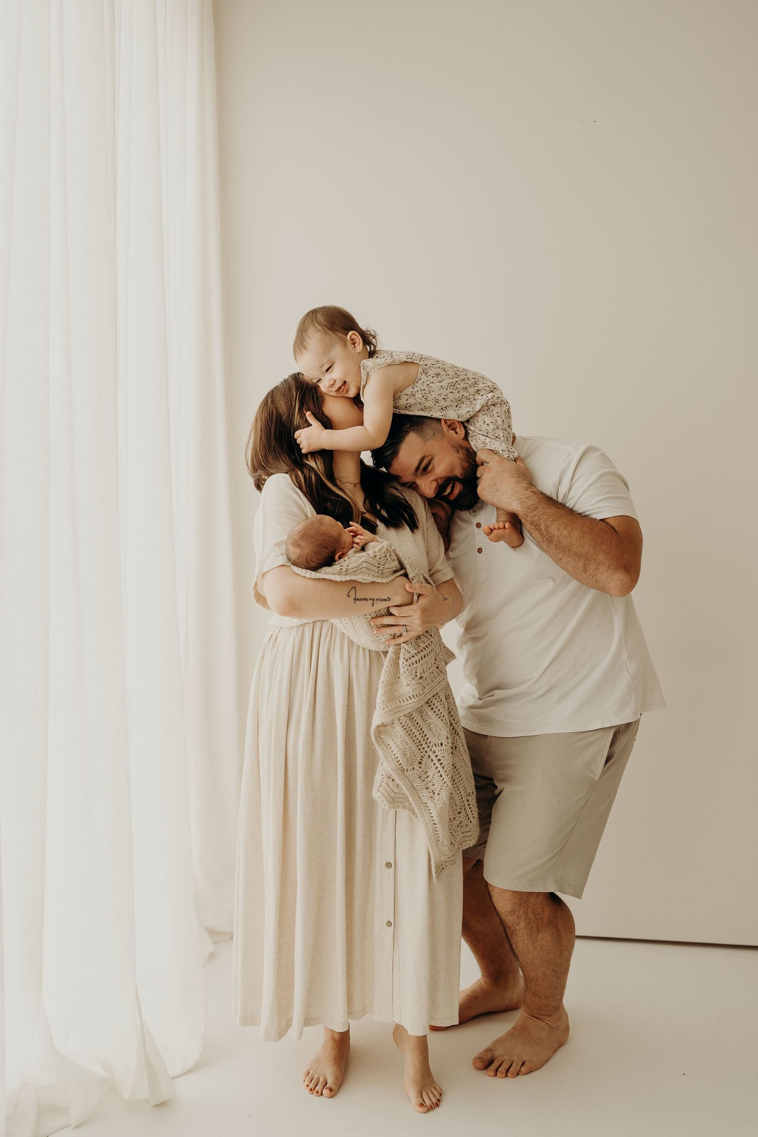 A joyful family portrait with parents holding a newborn and toddler in a bright, minimalist studio setting.