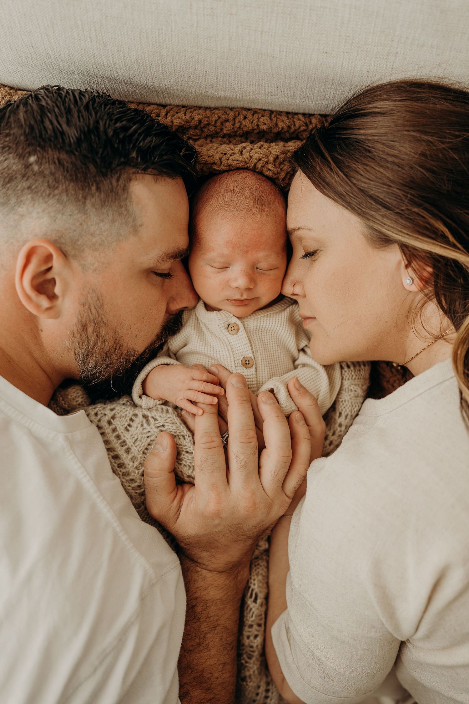 Two parents lying down, gently kissing their sleeping newborn baby dressed in a white onesie.