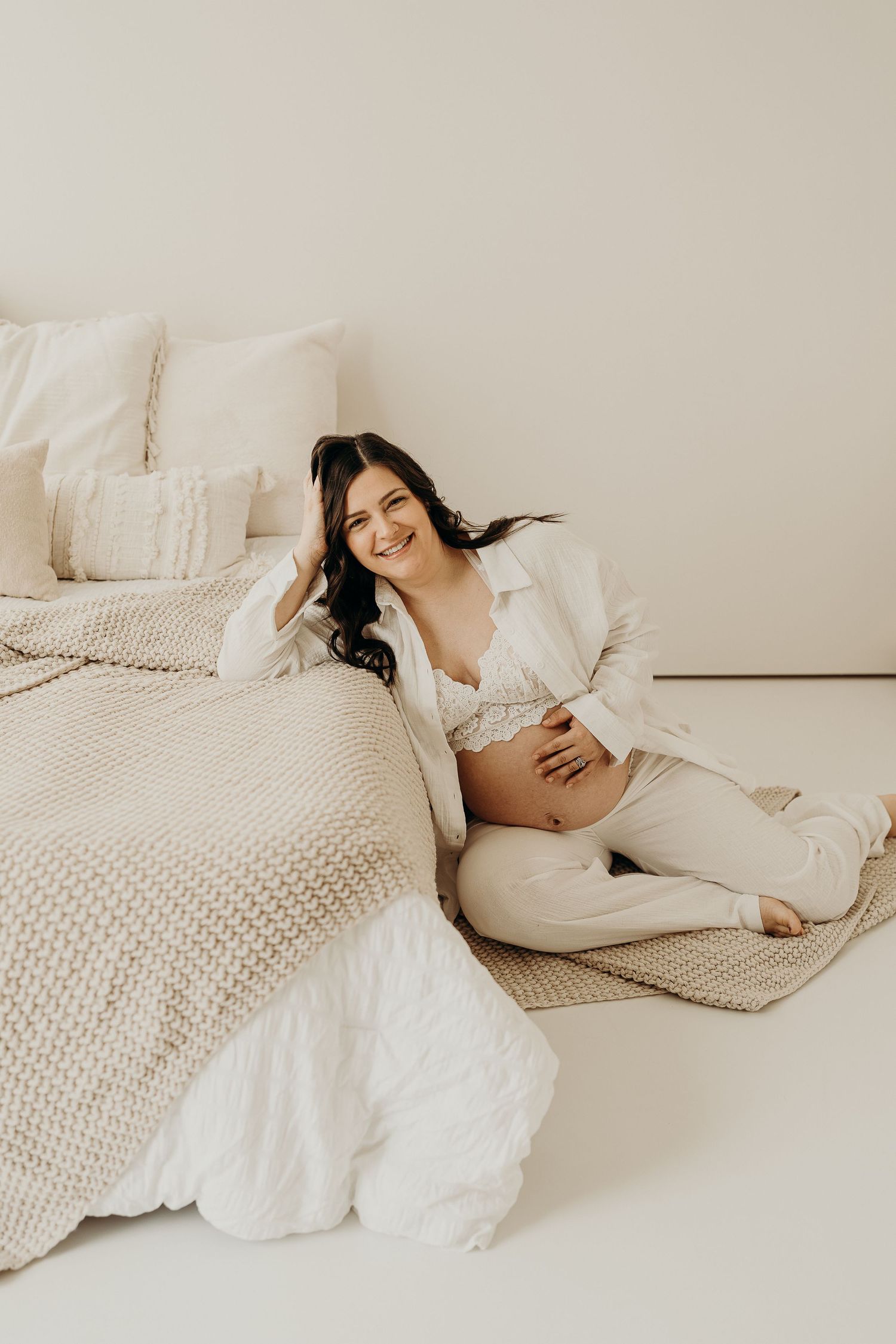 Smiling pregnant woman in beige outfit sitting on floor beside a bed, cradling her baby bump in a bright, neutral-toned room.