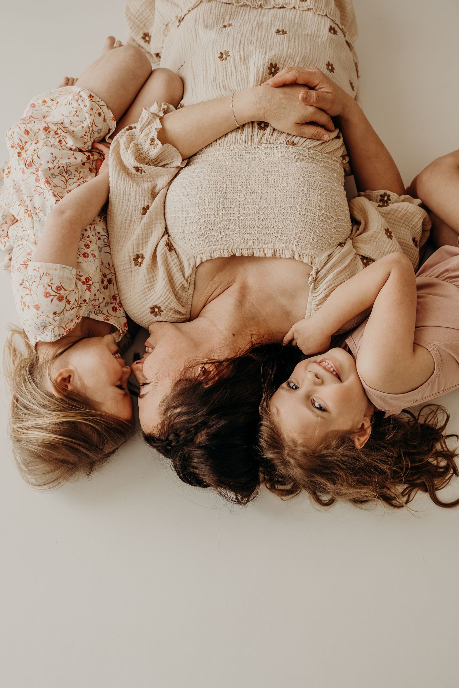 Pregnant woman lying down with two young daughters in floral dresses, sharing a tender family moment.
