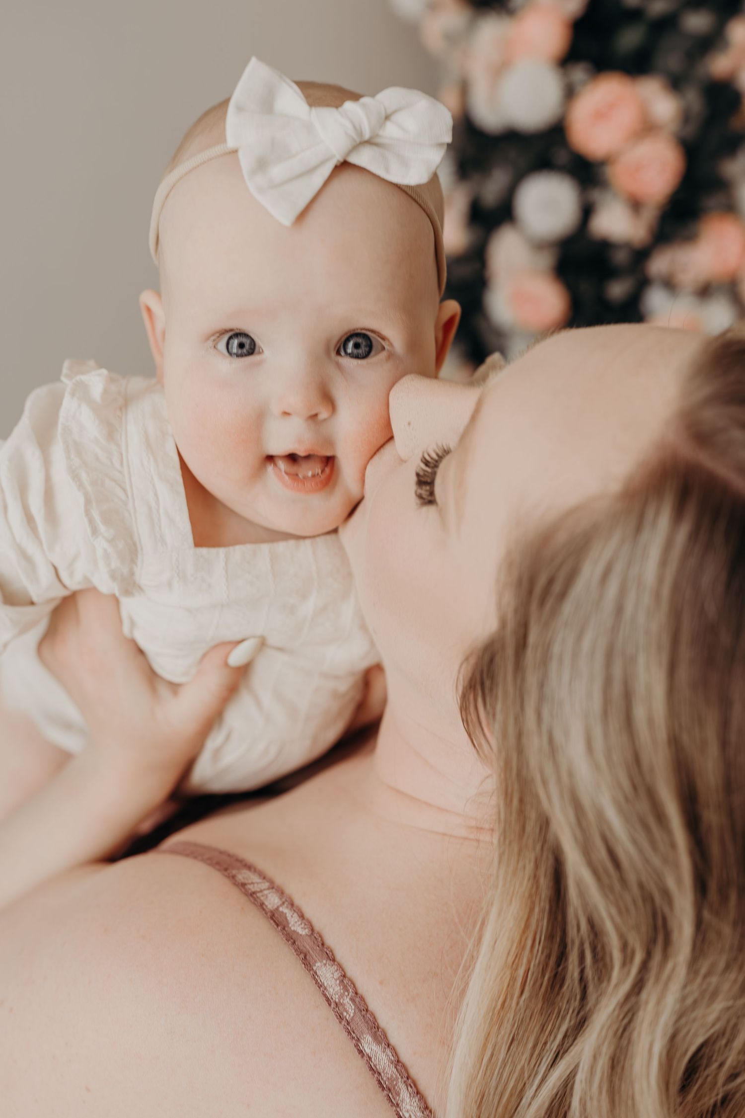 A mother tenderly kisses her smiling baby girl wearing a white bow headband and dress near floral decor during a motherhood mini session.