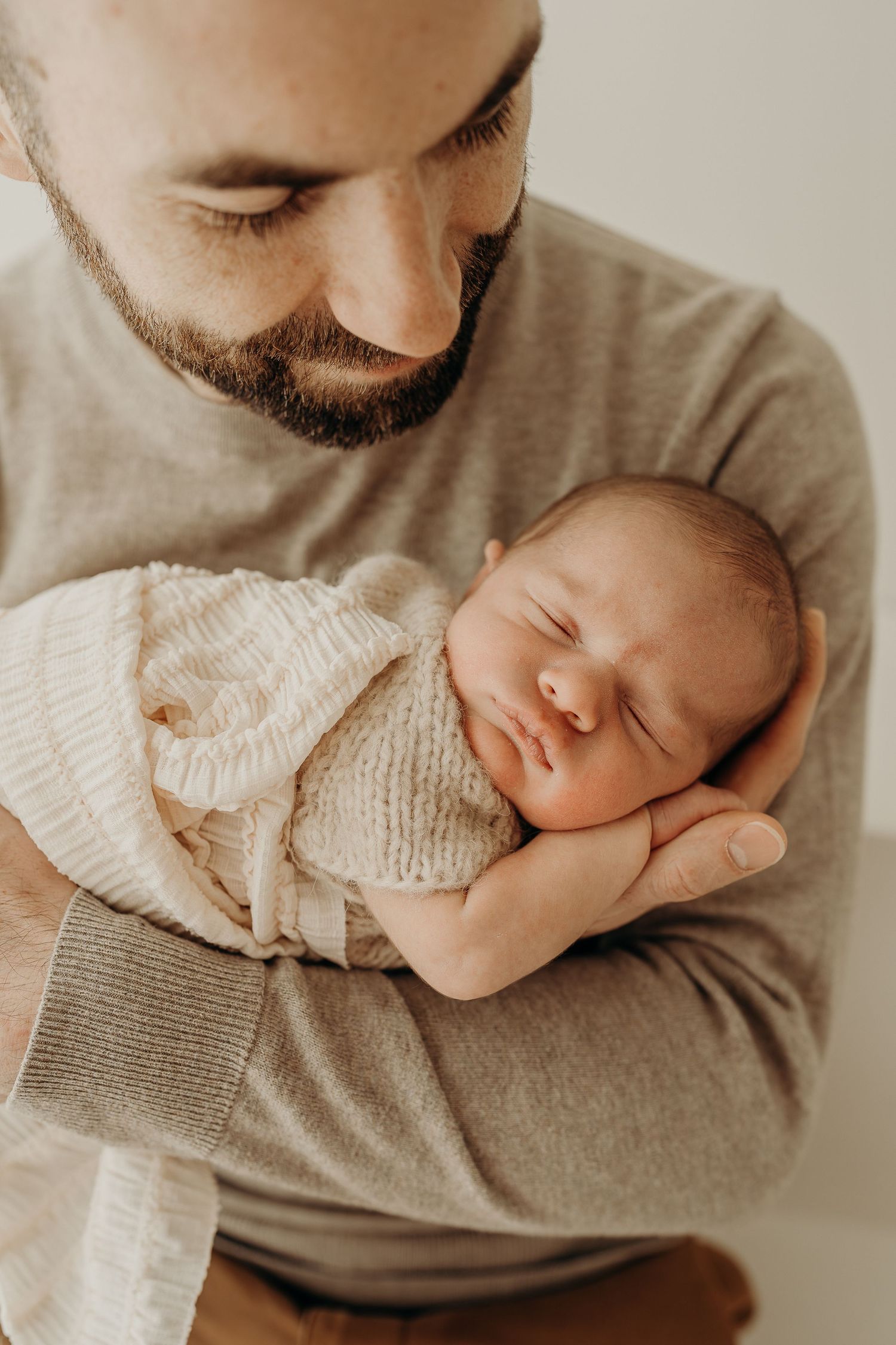 Bearded father tenderly holds sleeping newborn baby wrapped in cream knit blanket against his chest.