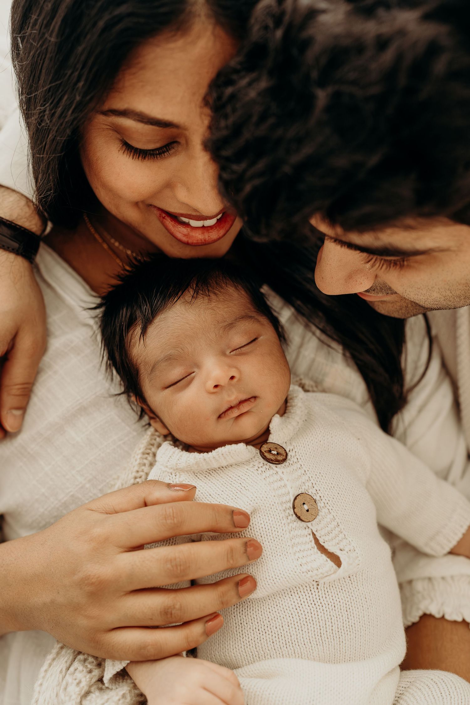 Parents lovingly gaze at their sleeping newborn baby dressed in a cream knit outfit during a family portrait session.