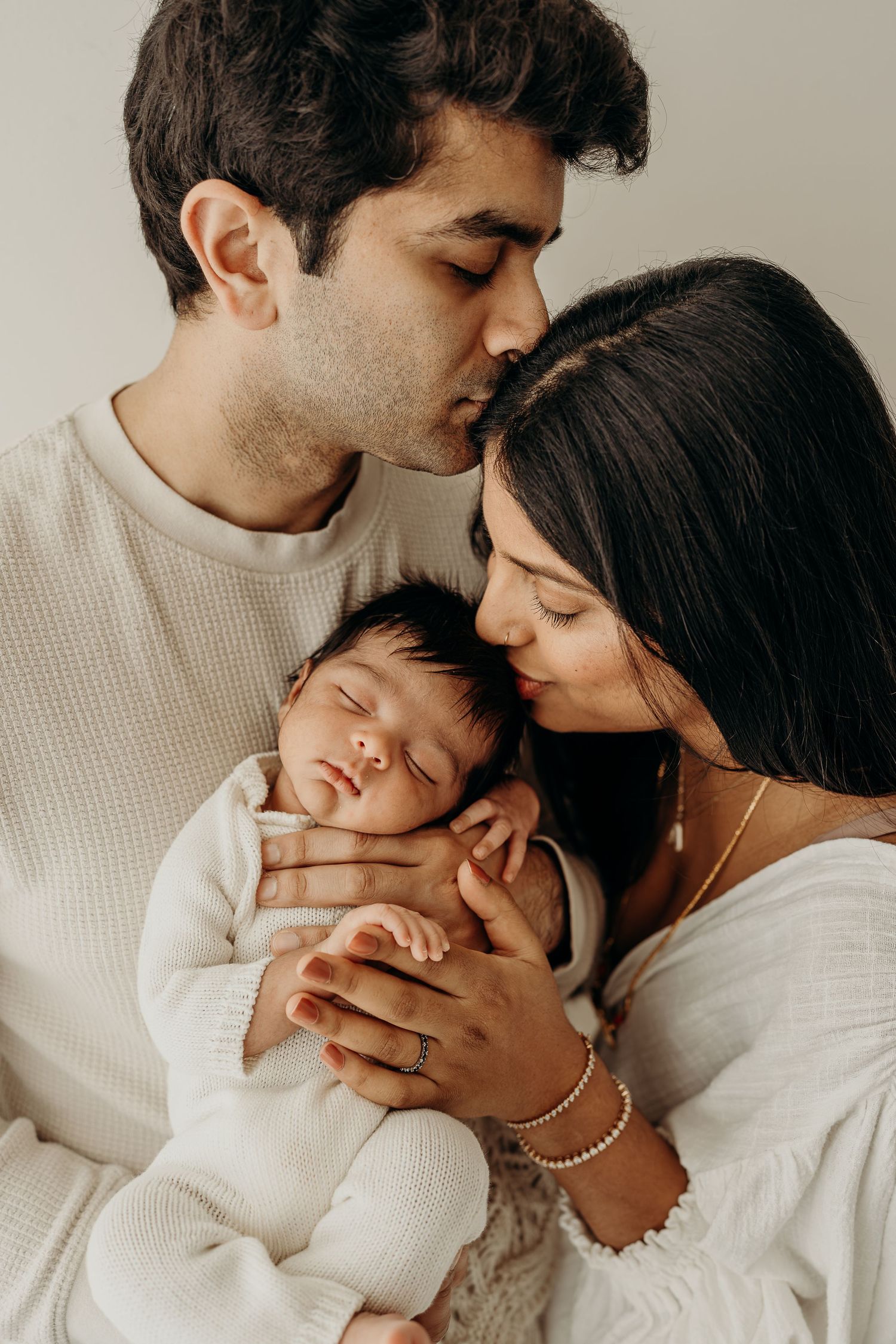 A couple in white sweaters tenderly kiss their sleeping newborn baby, capturing a serene family portrait.