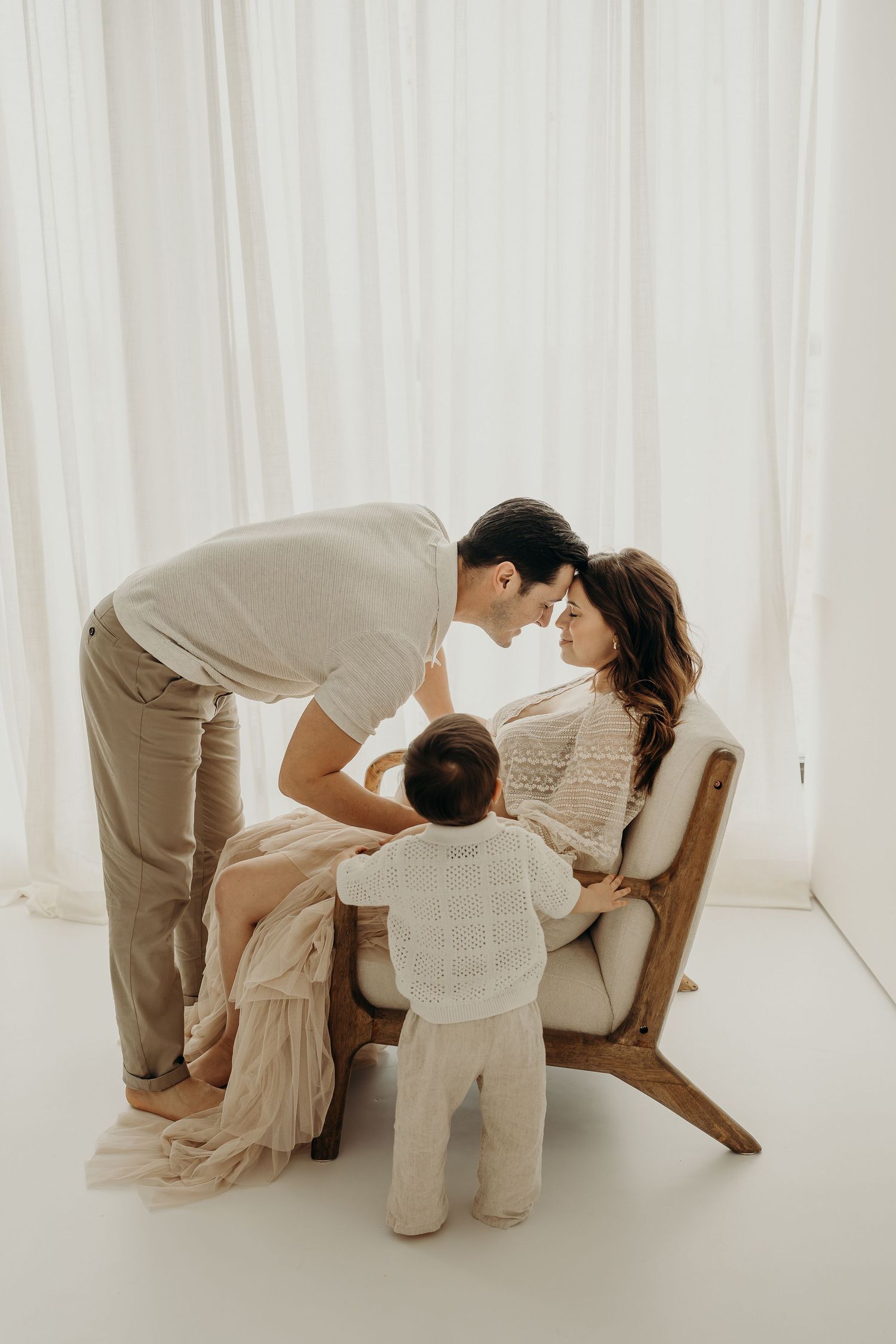 A loving family in neutral tones shares a tender moment, parents touching foreheads while toddler stands between them in a bright studio.