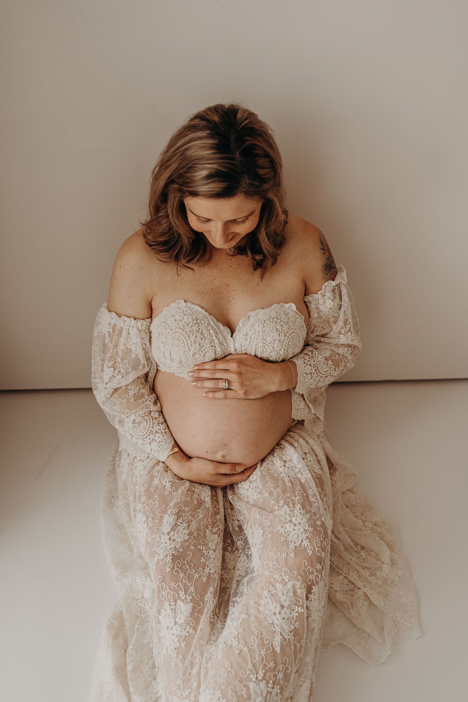 Pregnant woman in lace maternity gown cradling baby bump, elegant studio maternity photography session.