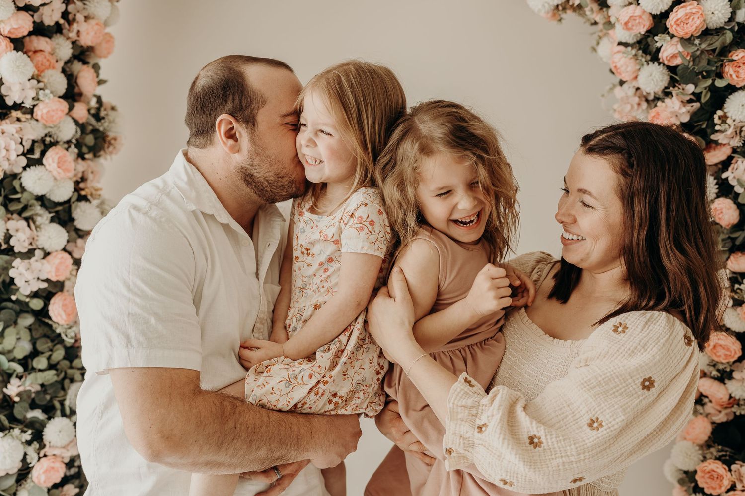 Happy family of four posing with pink floral arch backdrop, parents kissing and tickling two laughing little girls during a mini session.