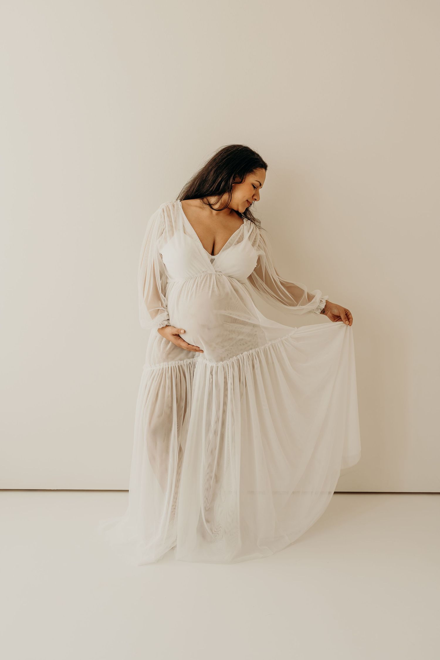 Ethereal maternity portrait in a flowing white dress against neutral background.
