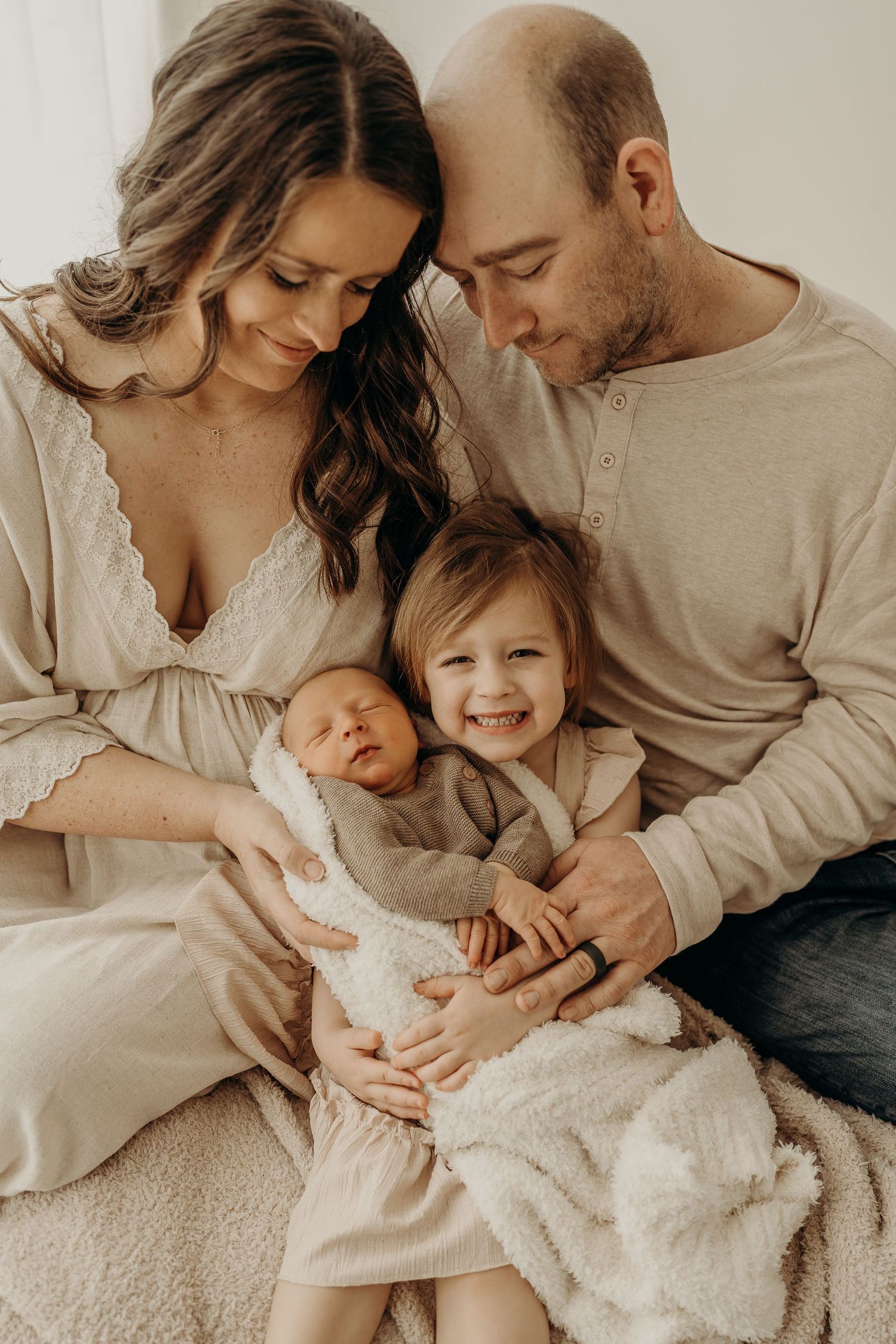 A happy family of four poses together, with parents holding a newborn baby and a smiling toddler girl in a warm, cozy setting.