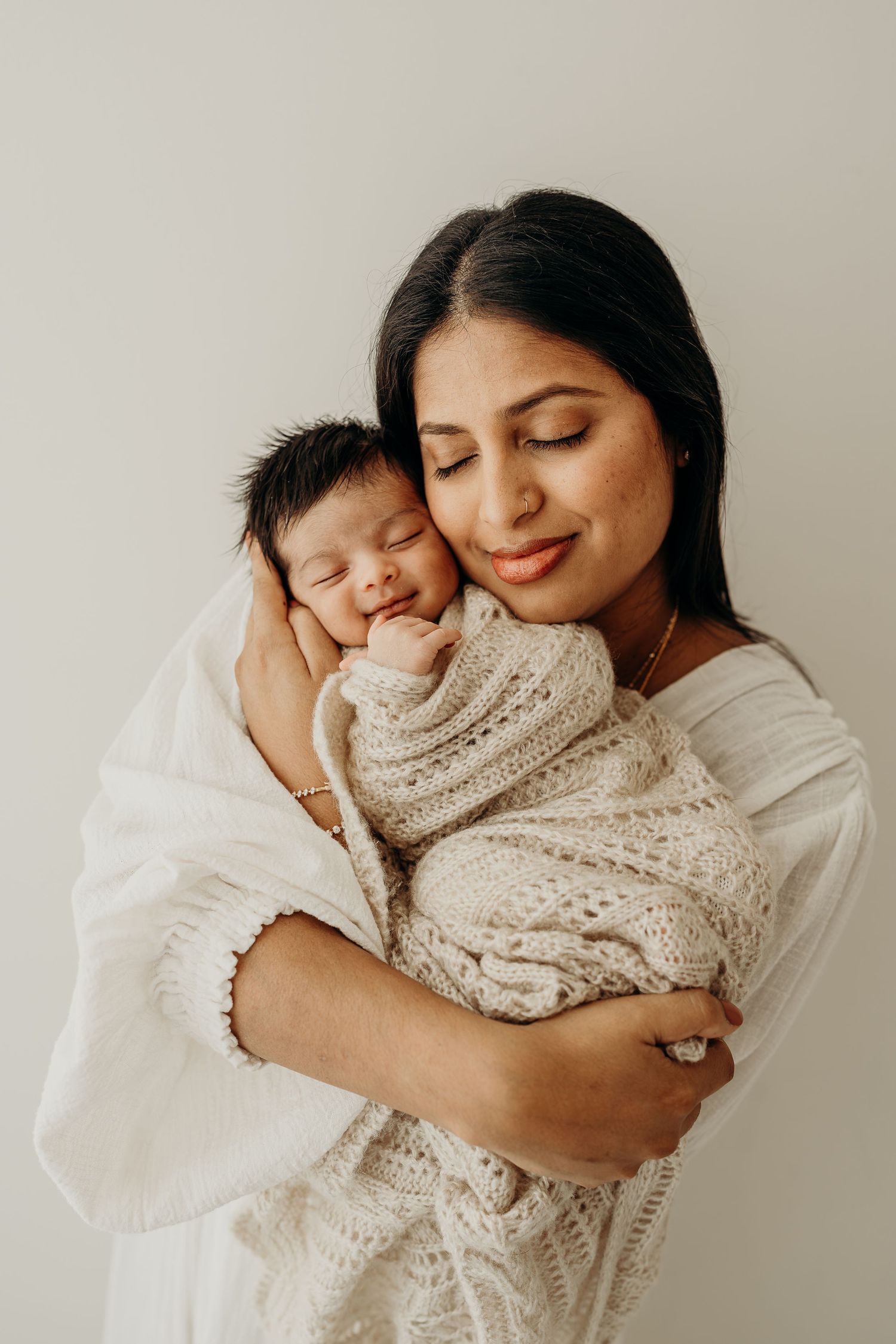 A mother tenderly holds her sleeping newborn wrapped in a cream knit blanket, both sharing a peaceful moment.