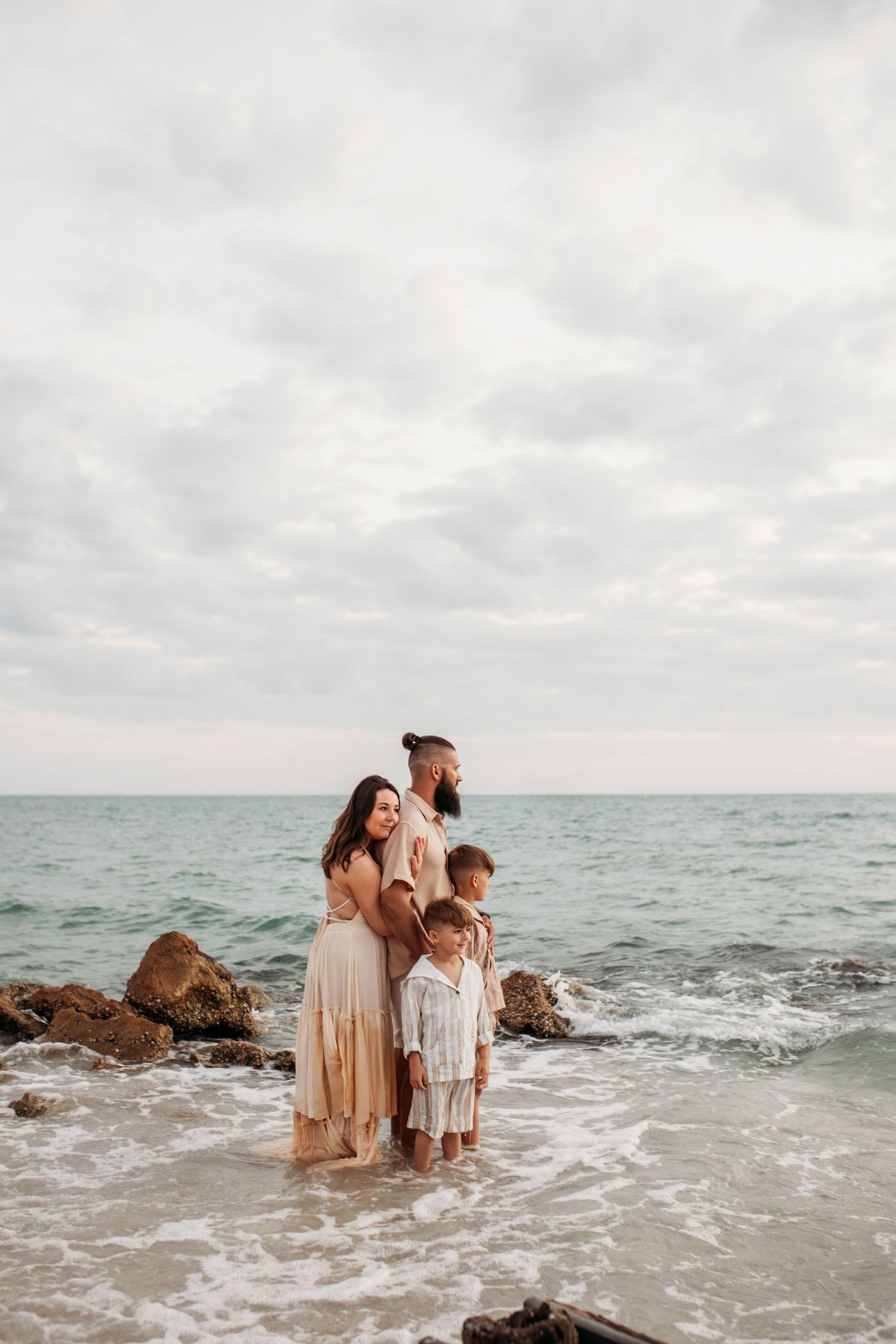 Family of four standing together in ocean waves on a rocky beach under a cloudy sky.