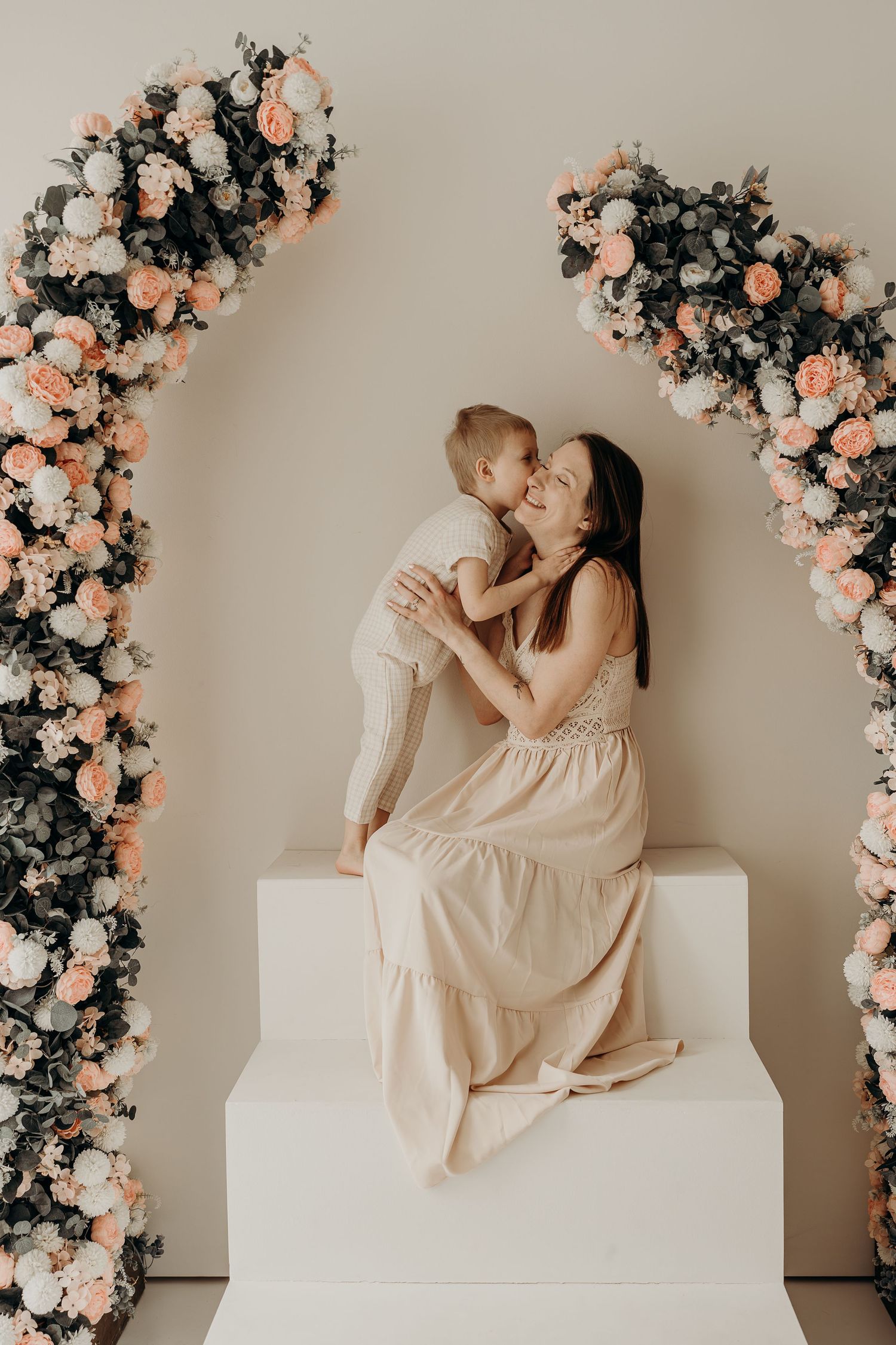 A mother in a dress sits on white steps kissing her young son, framed by floral arches with peach and white roses during a mini session.