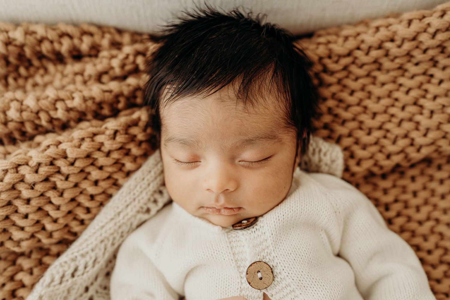 Peaceful newborn baby with dark hair sleeping on a chunky knit brown blanket wearing a white knit button cardigan.