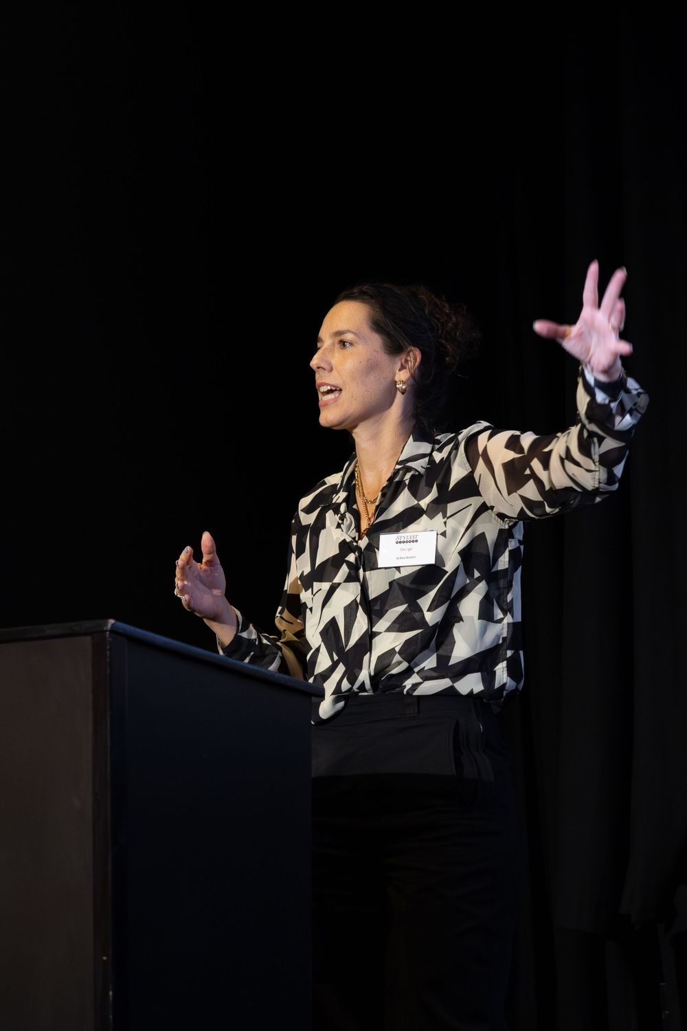 A speaker in a black and white patterned jacket gestures energetically while presenting at a podium on a dark stage.