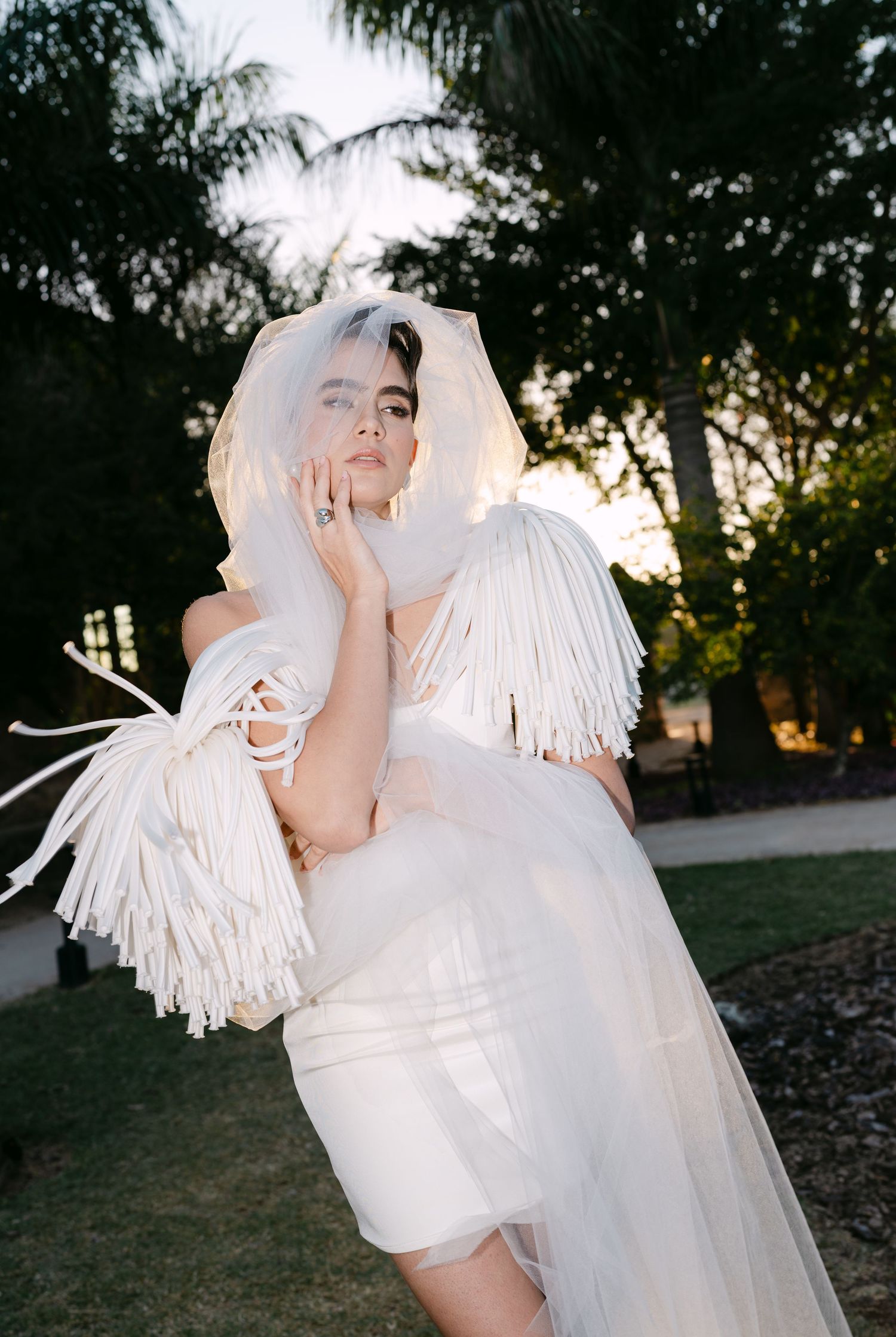 Bride in white fringe cape and veil poses pensively in lush garden at golden hour sunset.