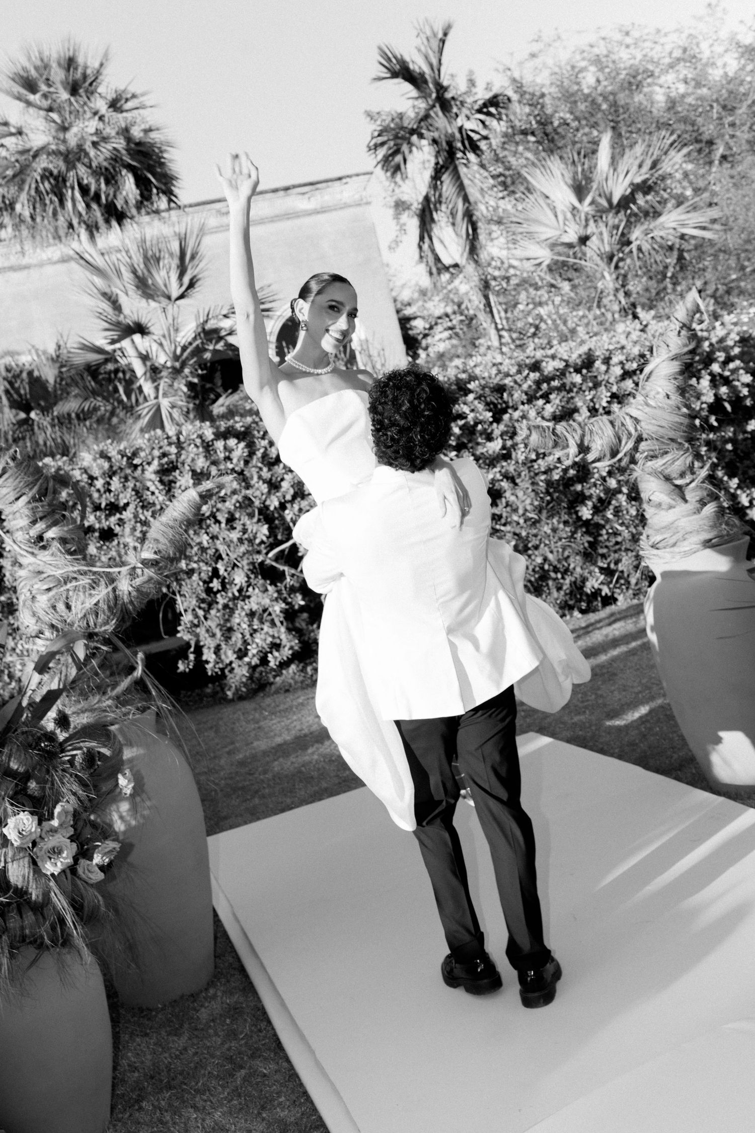 Black and white photo of groom lifting bride with arms raised joyfully at outdoor wedding reception.