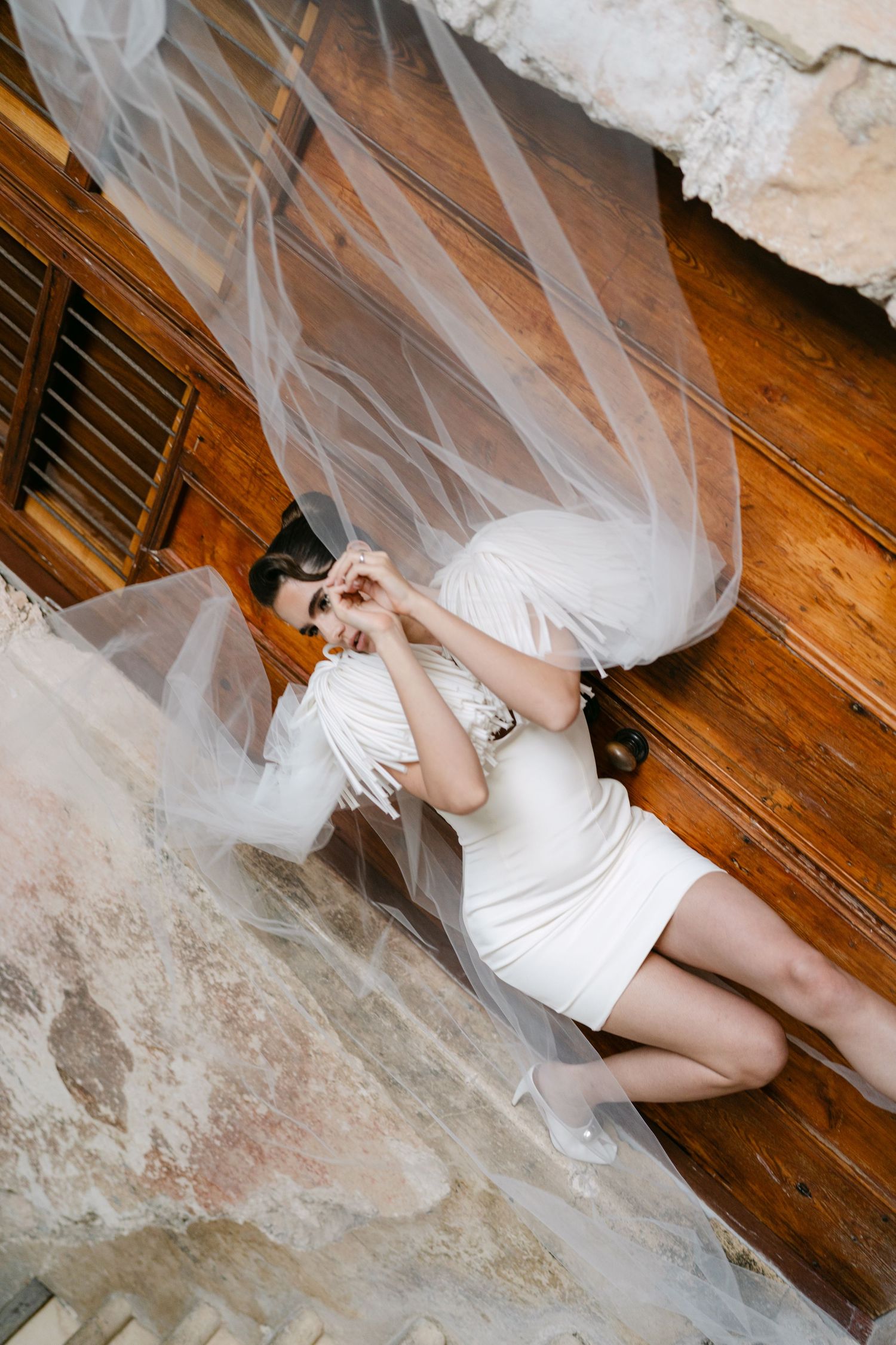 Bride in white mini dress lies on stone steps surrounded by flowing tulle veil.