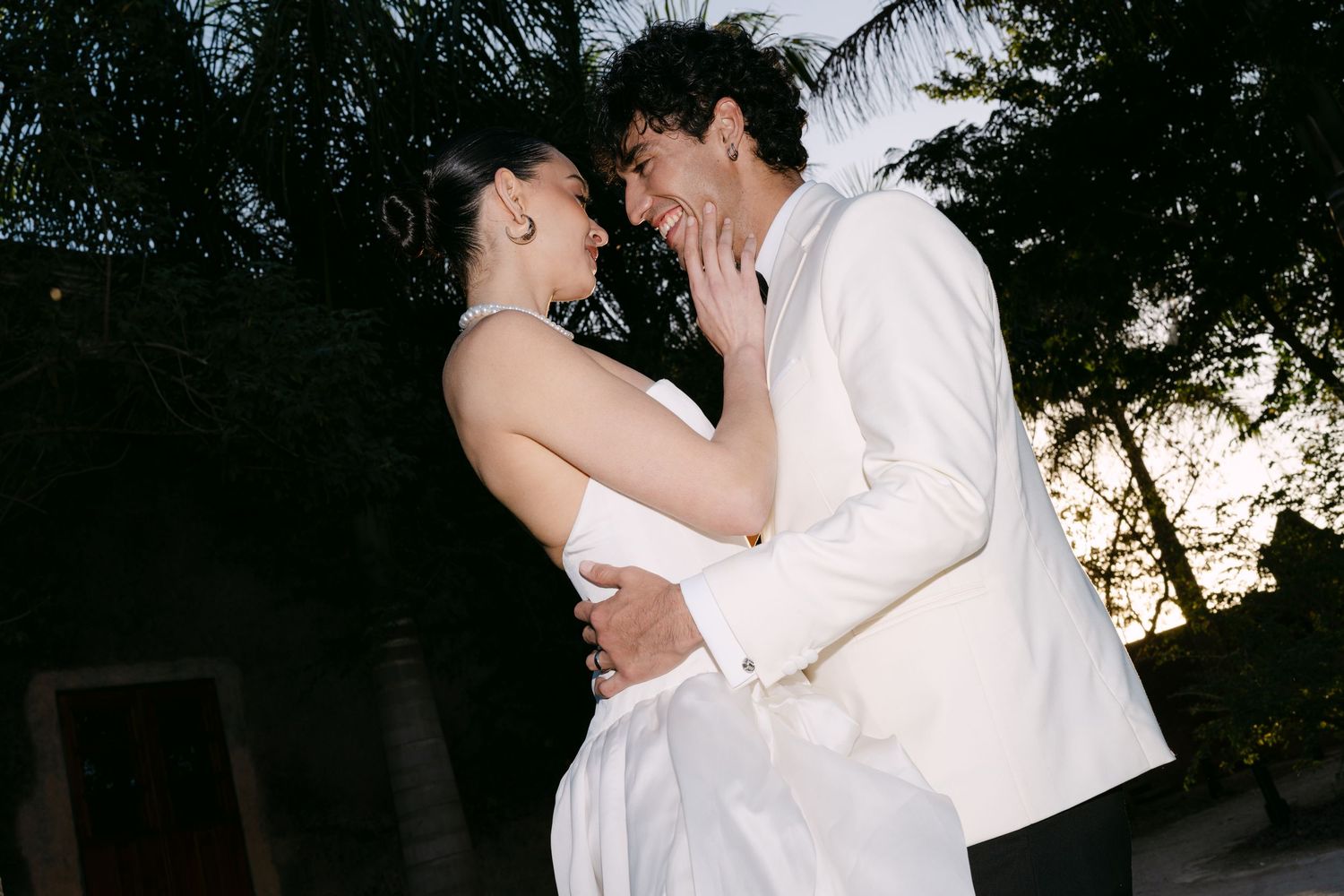 Bride and groom laugh and embrace intimately at dusk during their tropical outdoor wedding celebration.