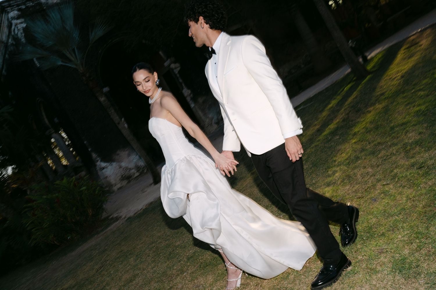 Bride in white gown and groom in white tuxedo jacket hold hands walking on grass at night.