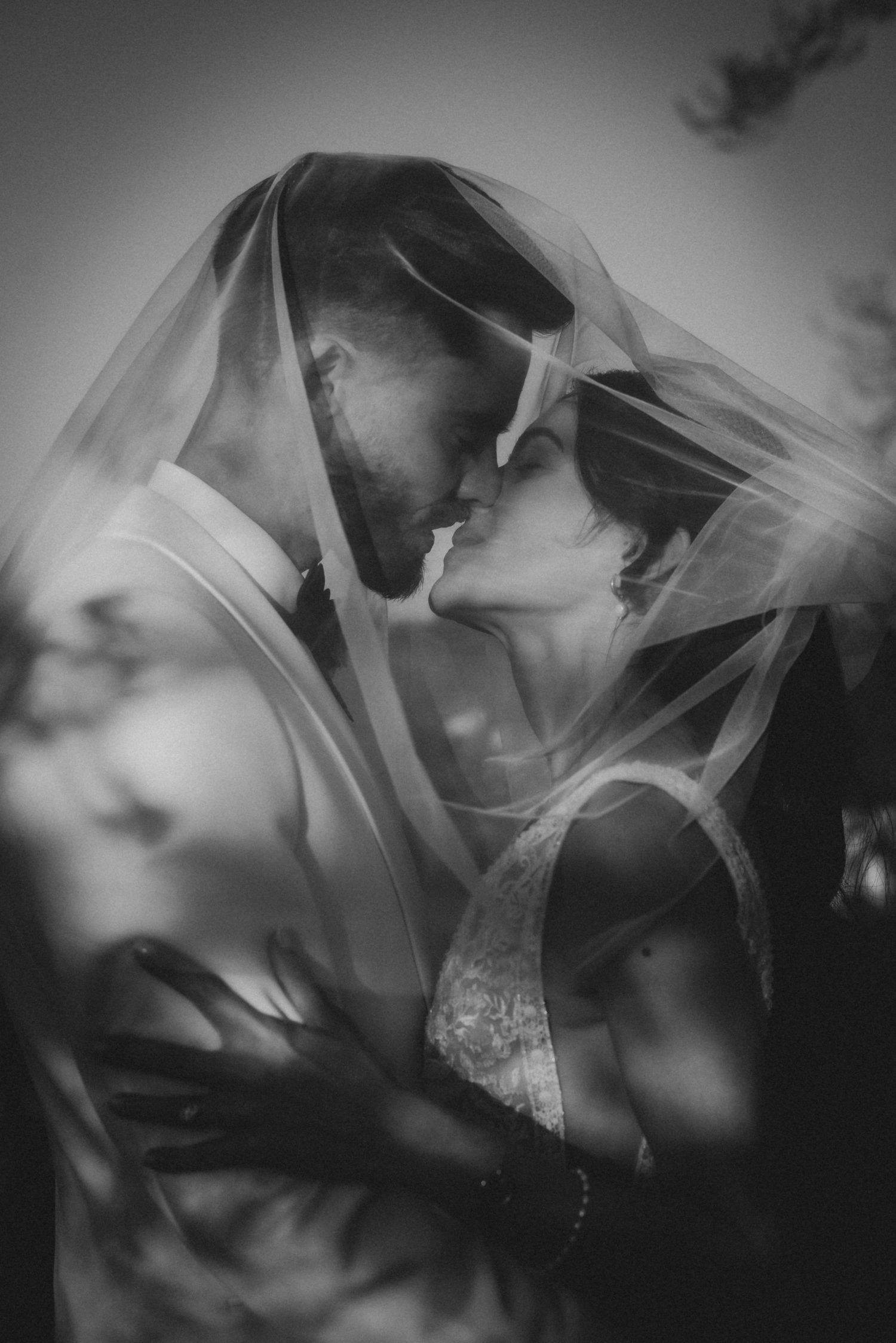 Black and white photo of a bride and groom sharing an intimate moment under a flowing veil.
