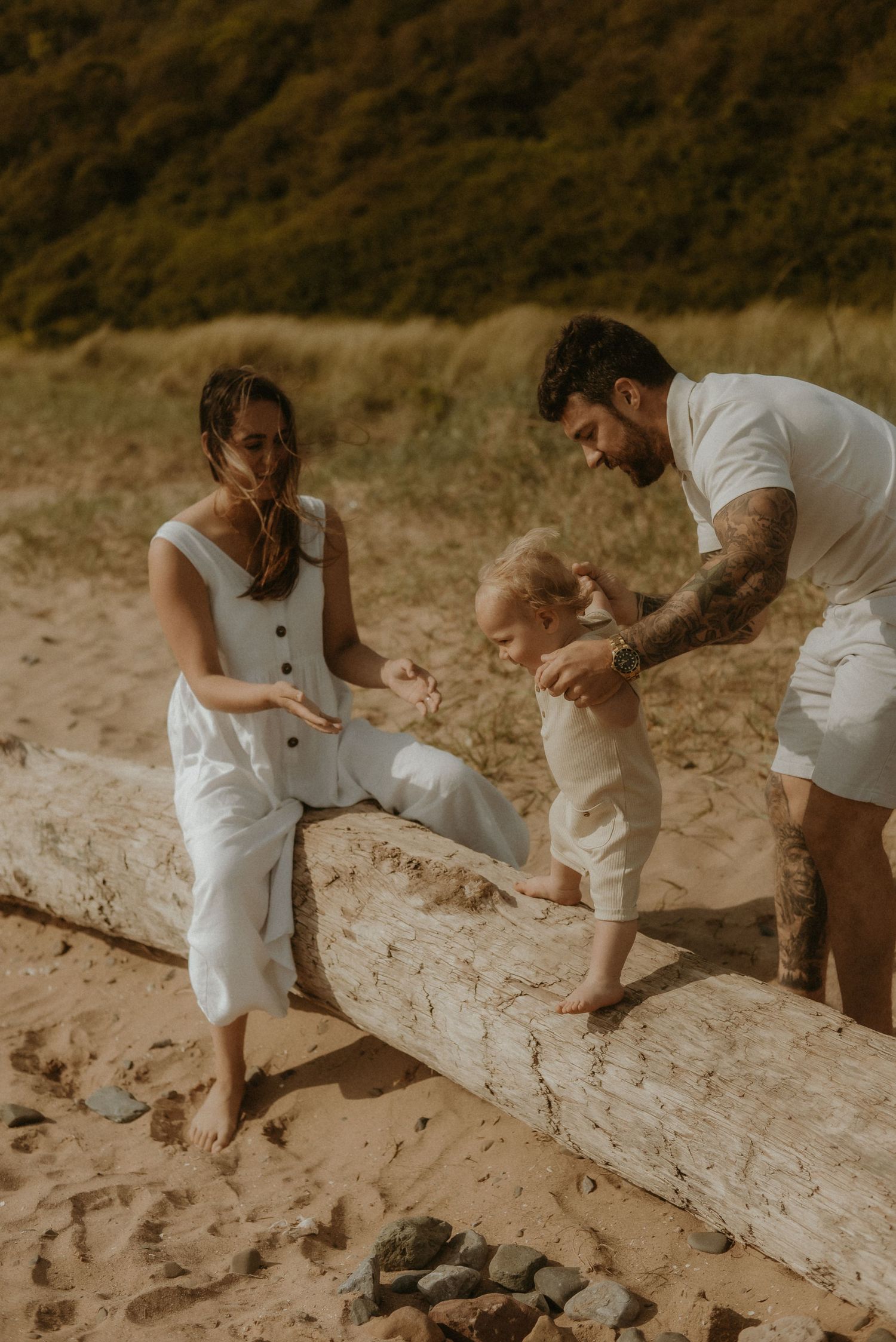 A young family in white outfits helps their toddler walk on driftwood at a sandy beach.