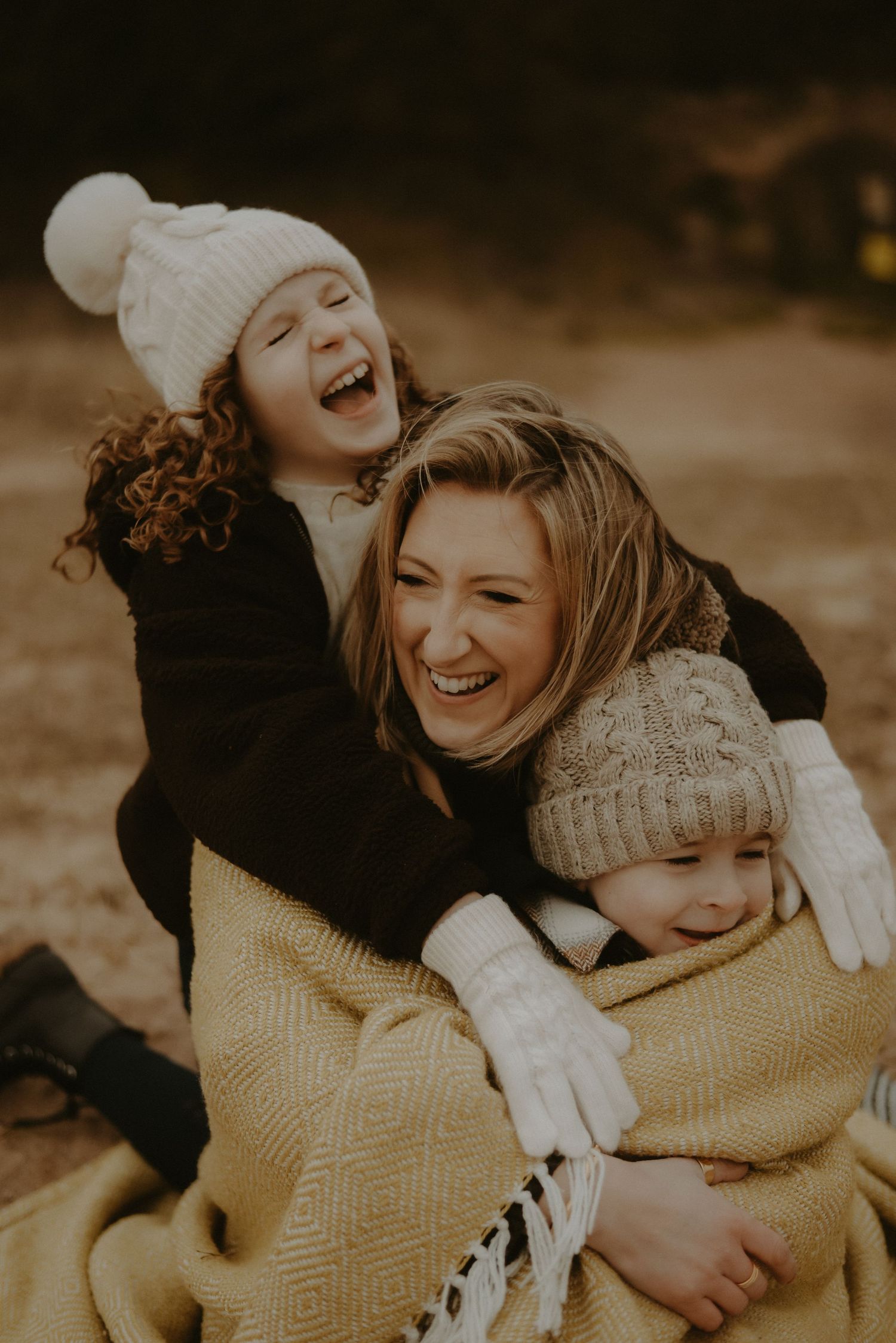 A mother and two children laughing joyfully wrapped in a yellow blanket wearing winter hats outdoors.