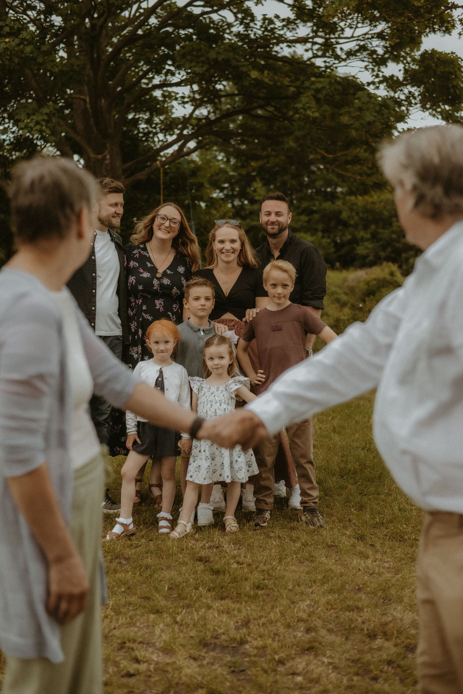 Large family group smiling outdoors on grass, with adults and children posing together near trees on a sunny day.