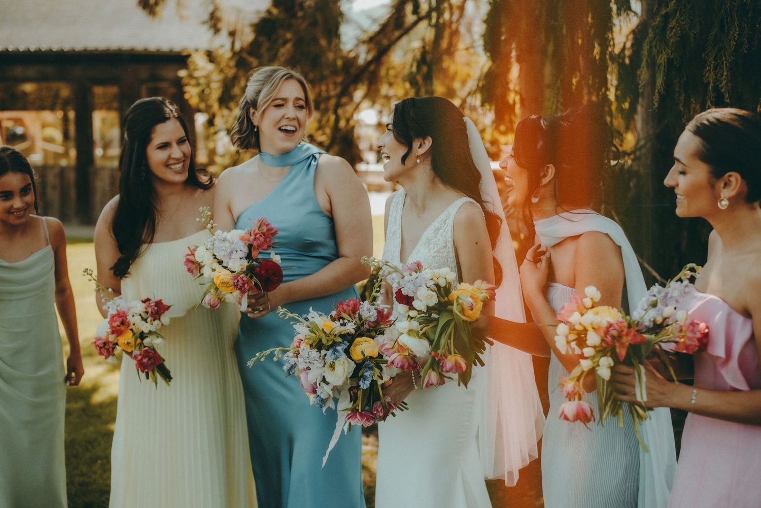 Bridesmaids in pastel dresses laughing and holding colorful floral bouquets at an outdoor wedding ceremony.