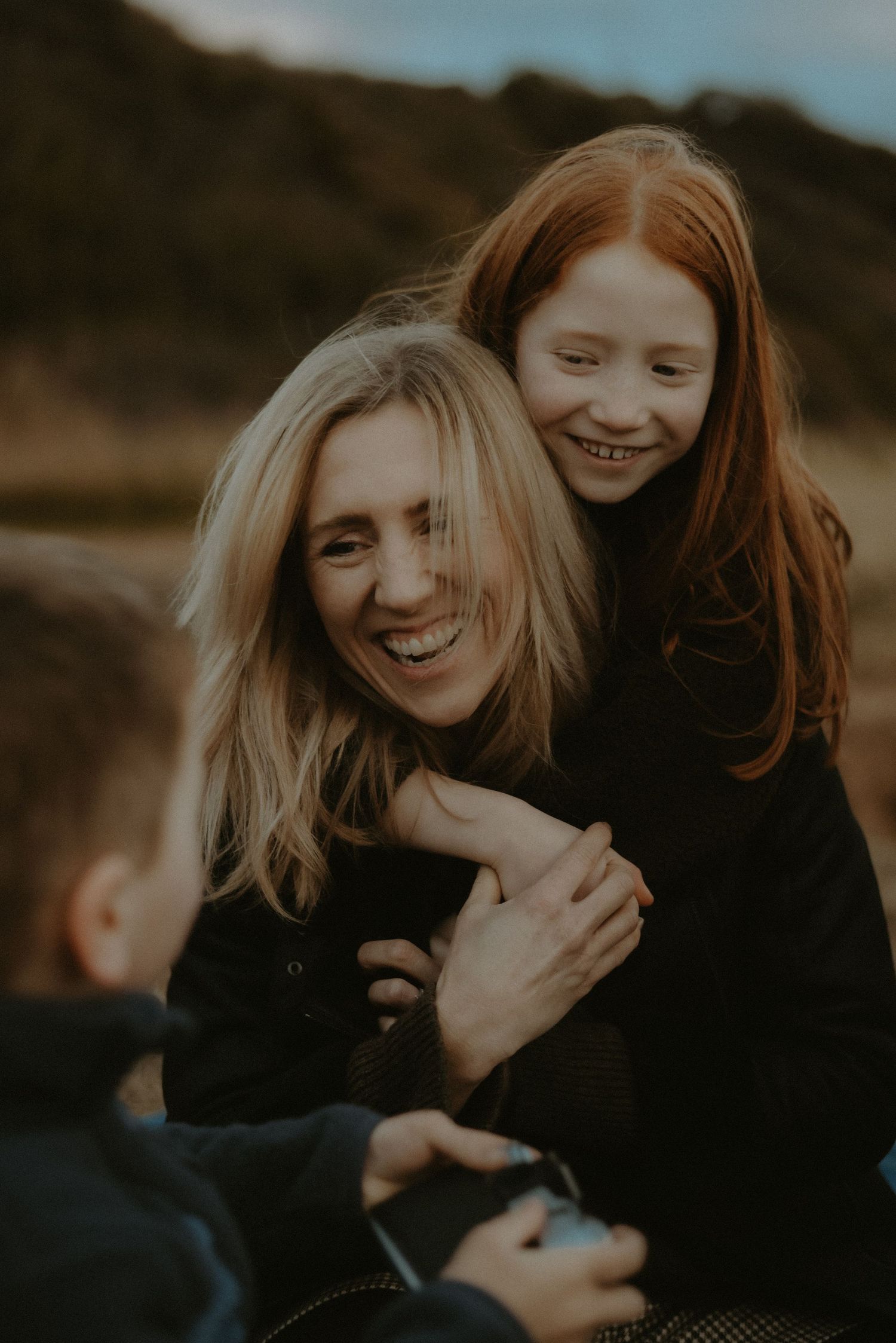 Laughing blonde mother and red-haired daughter sharing a joyful embrace outdoors in natural light.